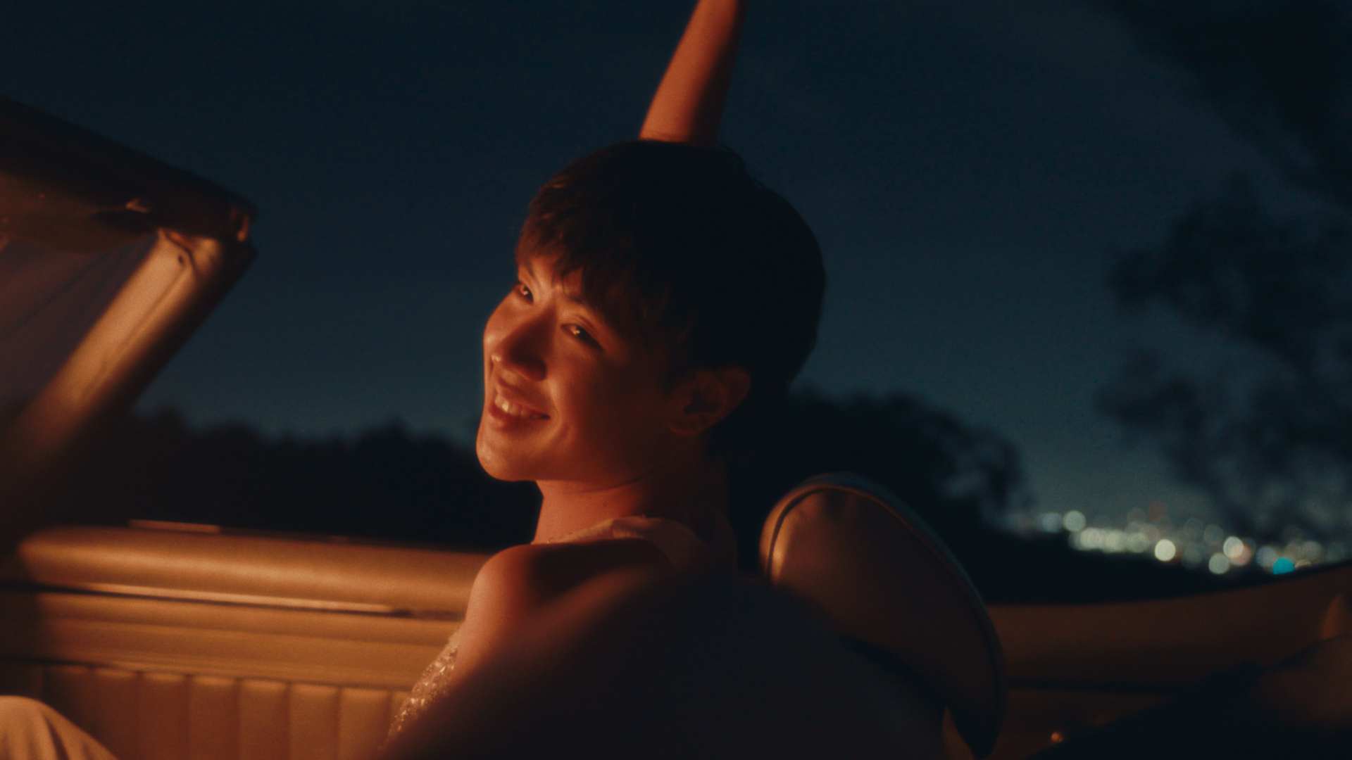 A person with short dark hair sitting inside a car at night, smiling and looking towards the camera, with city lights visible in the distance.