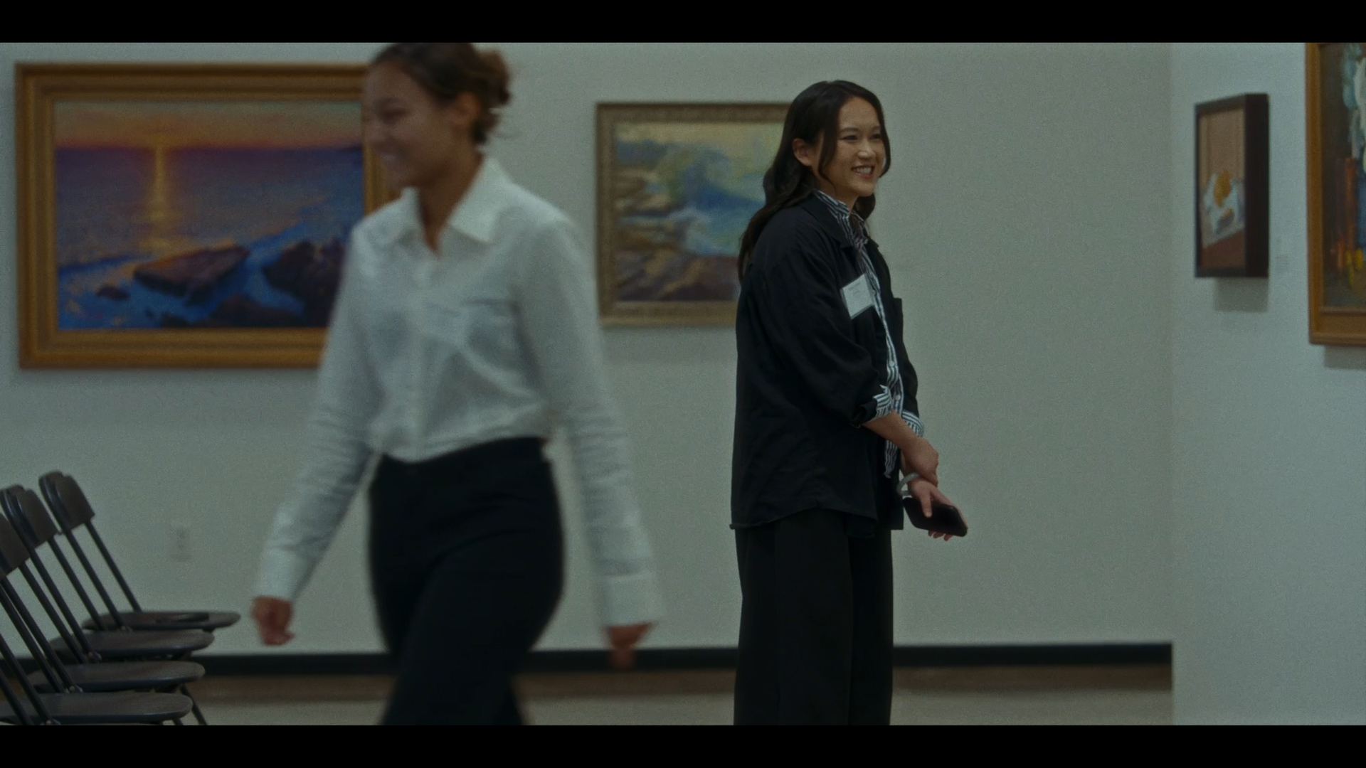 Two women in an art gallery, with paintings on the wall behind them. One woman is walking past, smiling, and the other is standing, dressed in black, holding a phone, and smiling.