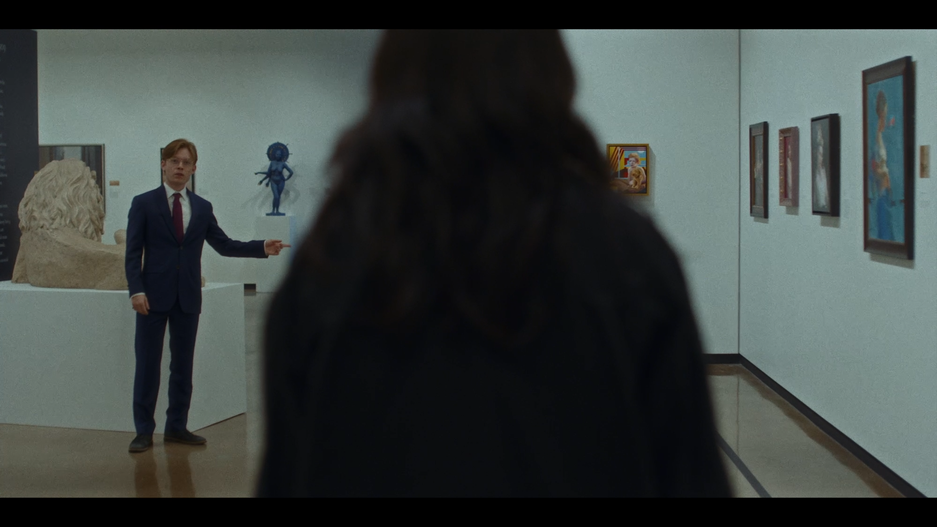 A person with long dark hair watches an art gallery exhibit featuring sculptures and paintings, with a male guide in a blue suit explaining. The gallery has white walls displaying various artworks.