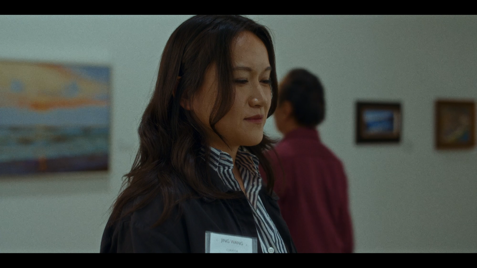 A woman with long wavy hair, wearing a dark jacket and striped shirt, is standing in front of a white wall with framed pictures, looking downward softly. Another person in the background is facing away, wearing a red shirt.