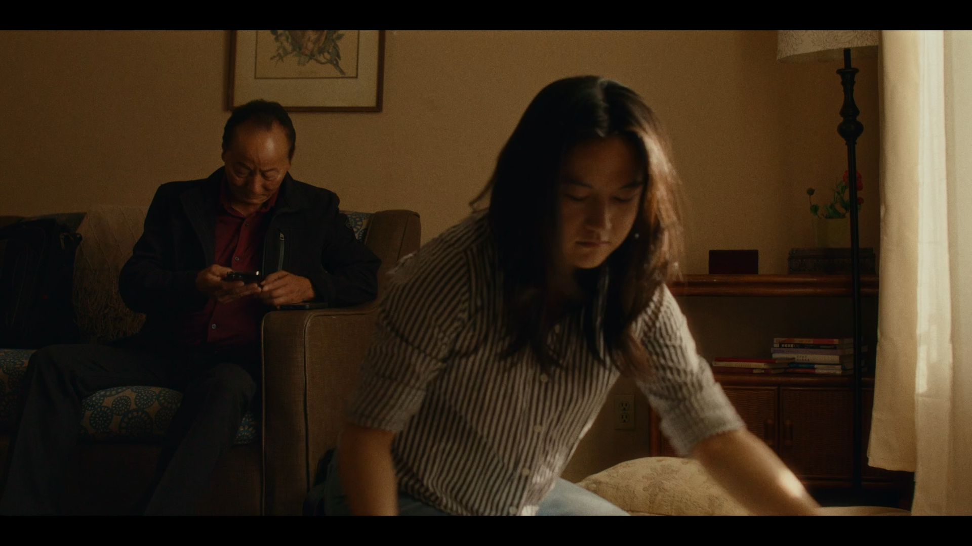 A man sitting on a couch looking at his phone, a woman in a striped shirt bending over, and a living room with a lamp, a bookshelf, and a window with curtains.