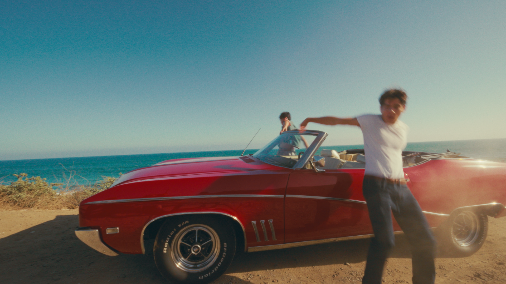 A person leaning on the front of a vintage red convertible car parked on a beachside with the ocean in the background, and another person standing in the car's driver seat, all during daytime with clear skies.