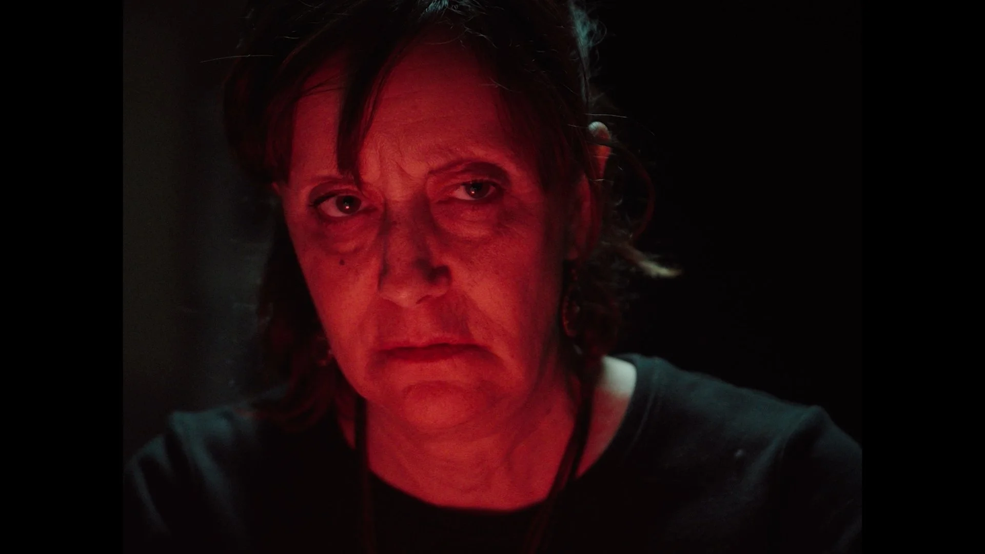A close-up portrait of a middle-aged woman with short, dark hair looking directly at the camera. The lighting is dim with a red hue, and the background is dark.