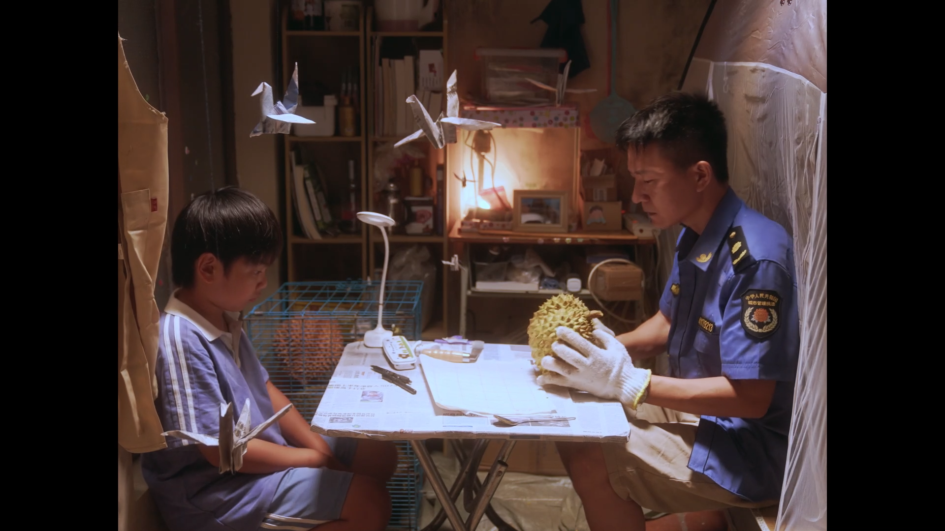 A boy sitting at a table with an older man in a uniform inspecting a durian fruit. The environment appears to be a small room with shelves and various objects in the background. The boy looks down at the table, while the man holds the durian and exam
