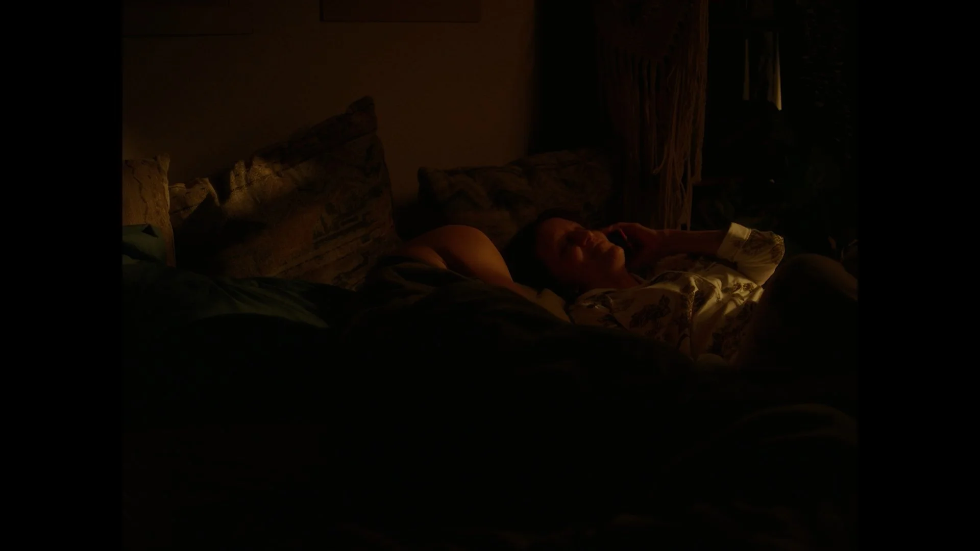 Person lying on bed in dark room, illuminated by soft light, with head resting on pillow, surrounded by patterned cushions.