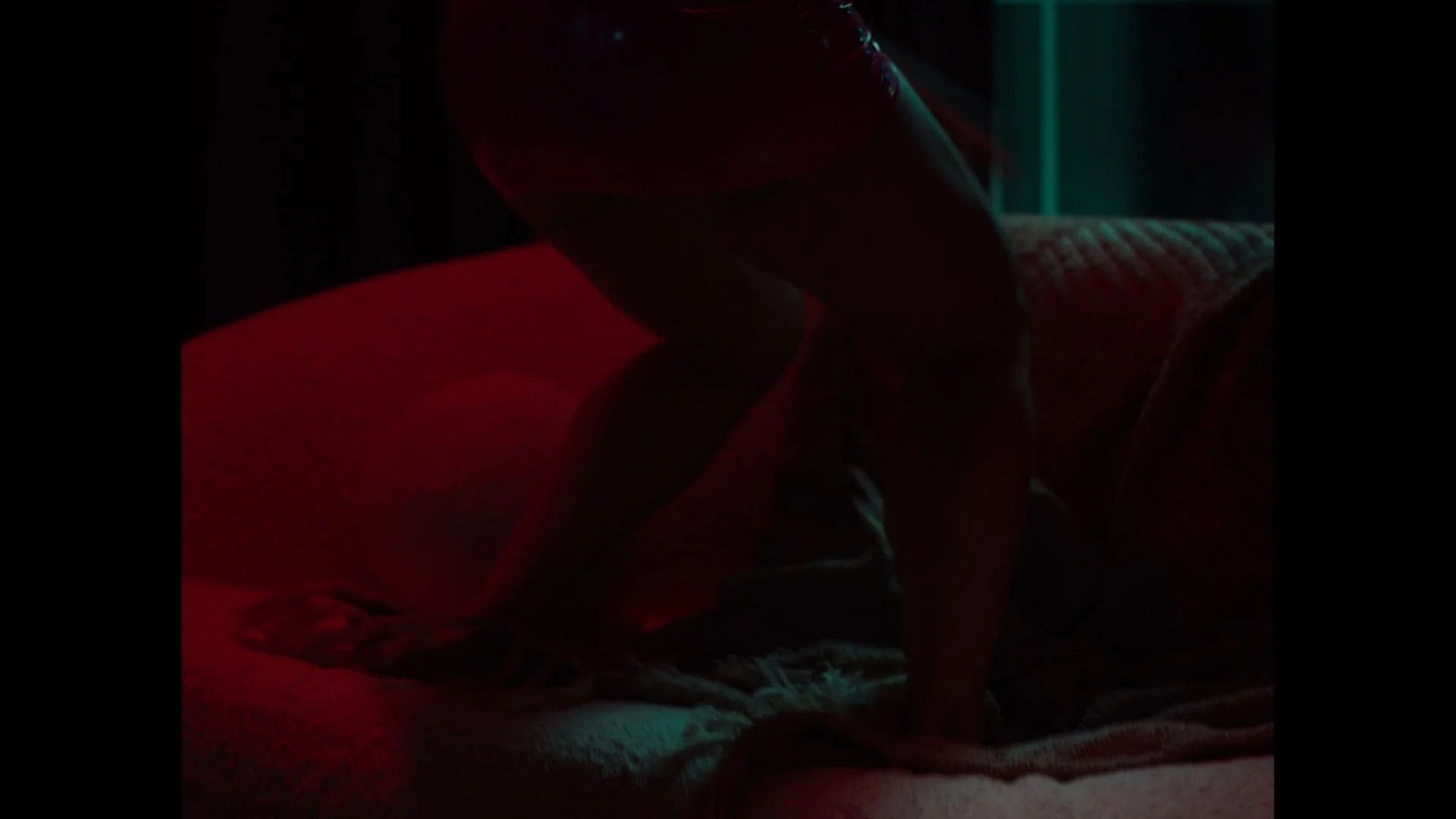 A person is on a bed in a dark room illuminated by red and teal lighting, with their body bent forward and arms on the bed.