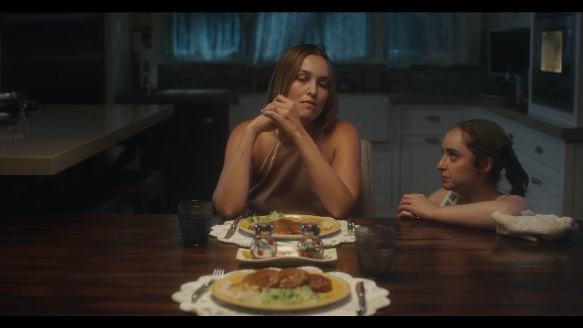A woman and a young girl sit at a dining table with plates of food, in a dimly lit kitchen, looking at each other.