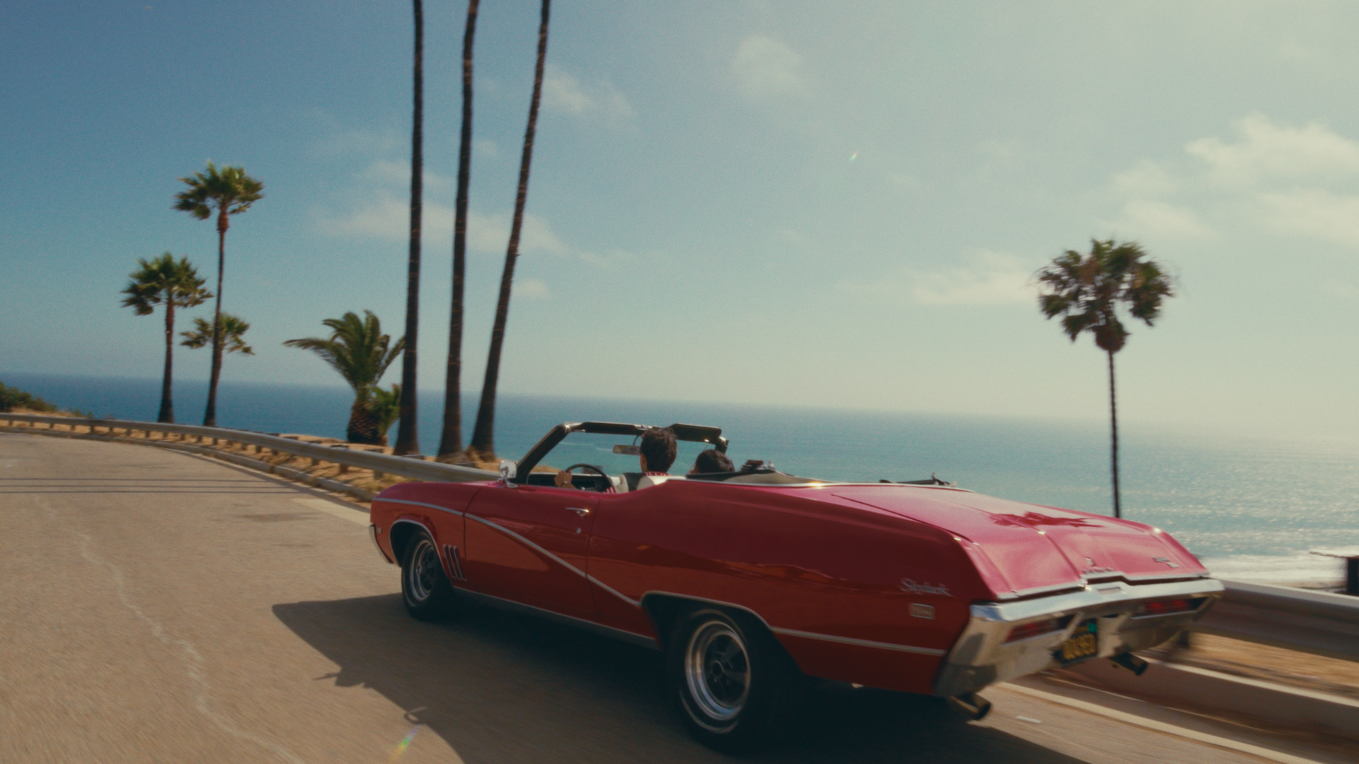 A red convertible car with two people inside driving along a coastal road with palm trees and the ocean in the background.