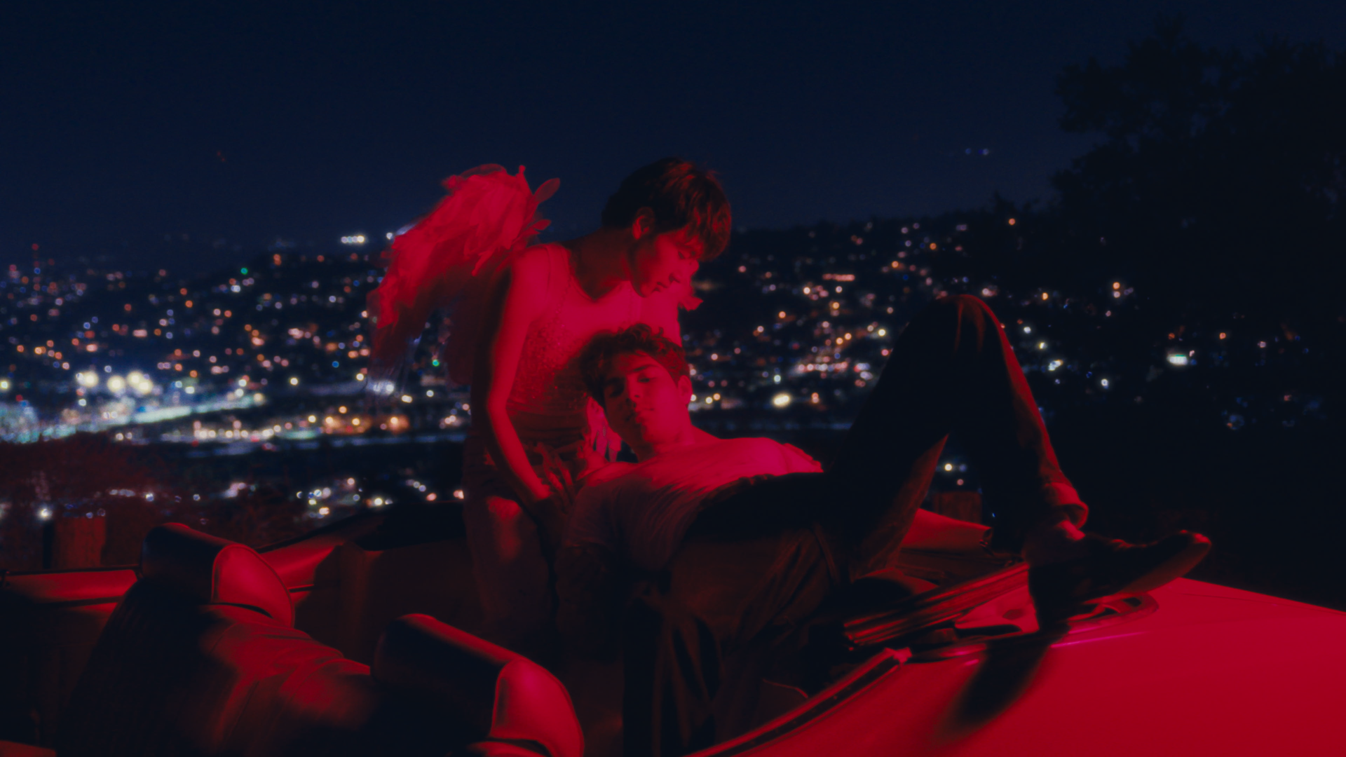 Two young men with angel wings leaning over a person lying on an inflatable boat at night, with a cityscape in the background illuminated by city lights.