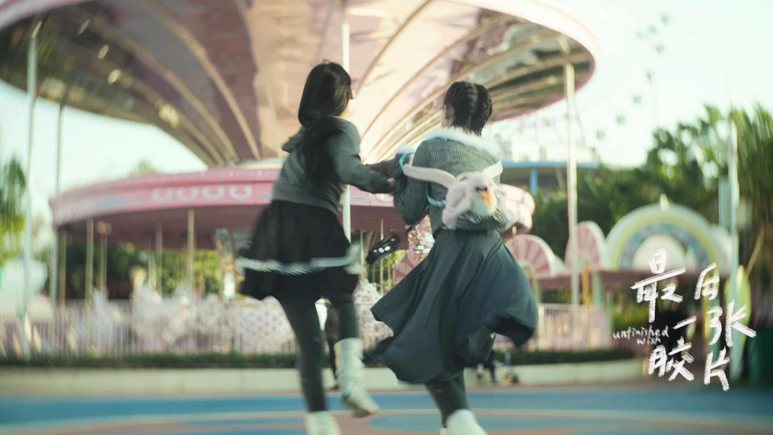 Two young women are running and holding hands on a colorful amusement park ride with a pink canopy and carousel horses in the background.