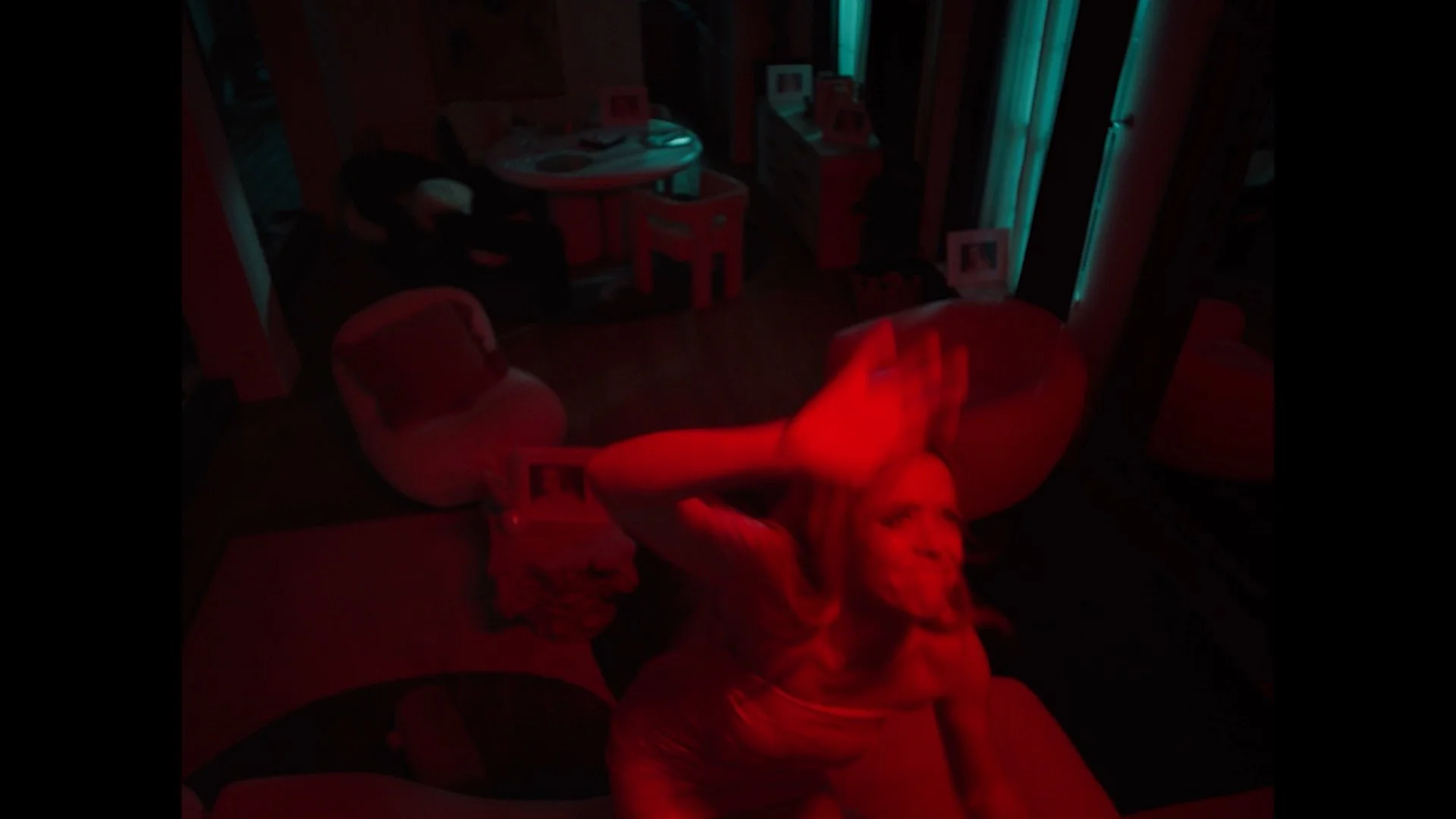 A woman taking a selfie in a dimly lit room illuminated by red and blue lights, with furniture including a bean bag chair, a small table, and picture frames visible in the background.