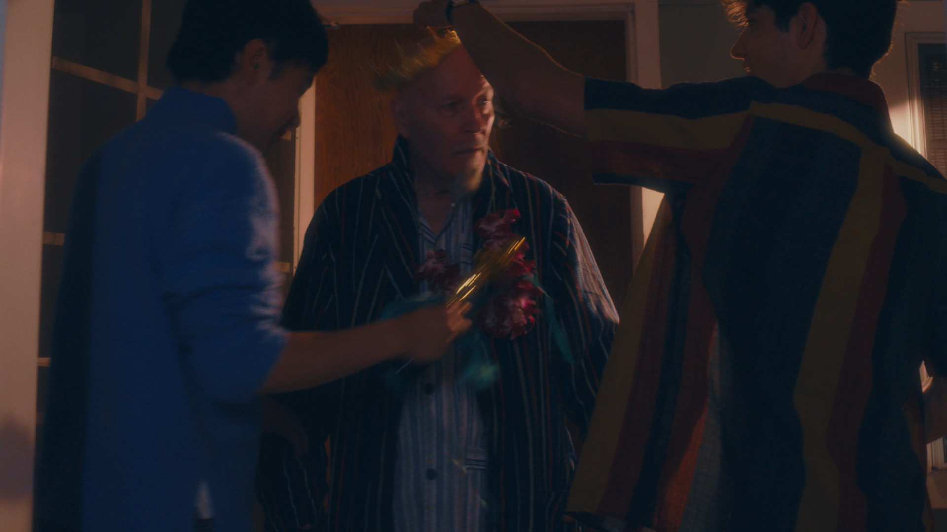 Three men in a dimly lit room celebrating a birthday. One man in the center wearing a striped shirt and a floral lei is having a birthday hat placed on his head. The man on the right wearing a striped sweater is smiling, and the man on the left is ho