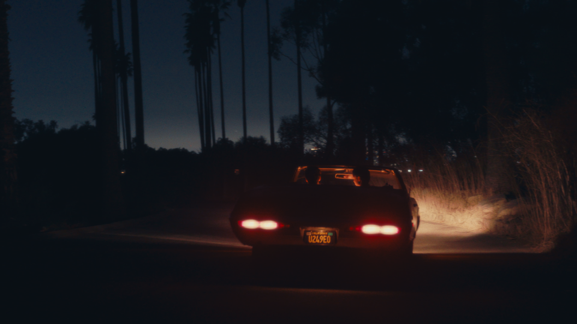 A vintage car driving on a dark, rural road at night, with tall trees silhouetted against the night sky and some distant city lights in the background.