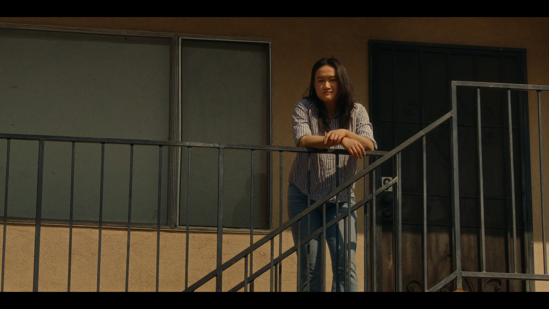 A woman standing on a balcony, leaning on the railing, with a neutral expression, wearing a striped shirt and jeans, in front of a beige building with sliding glass doors and a black metal gate.