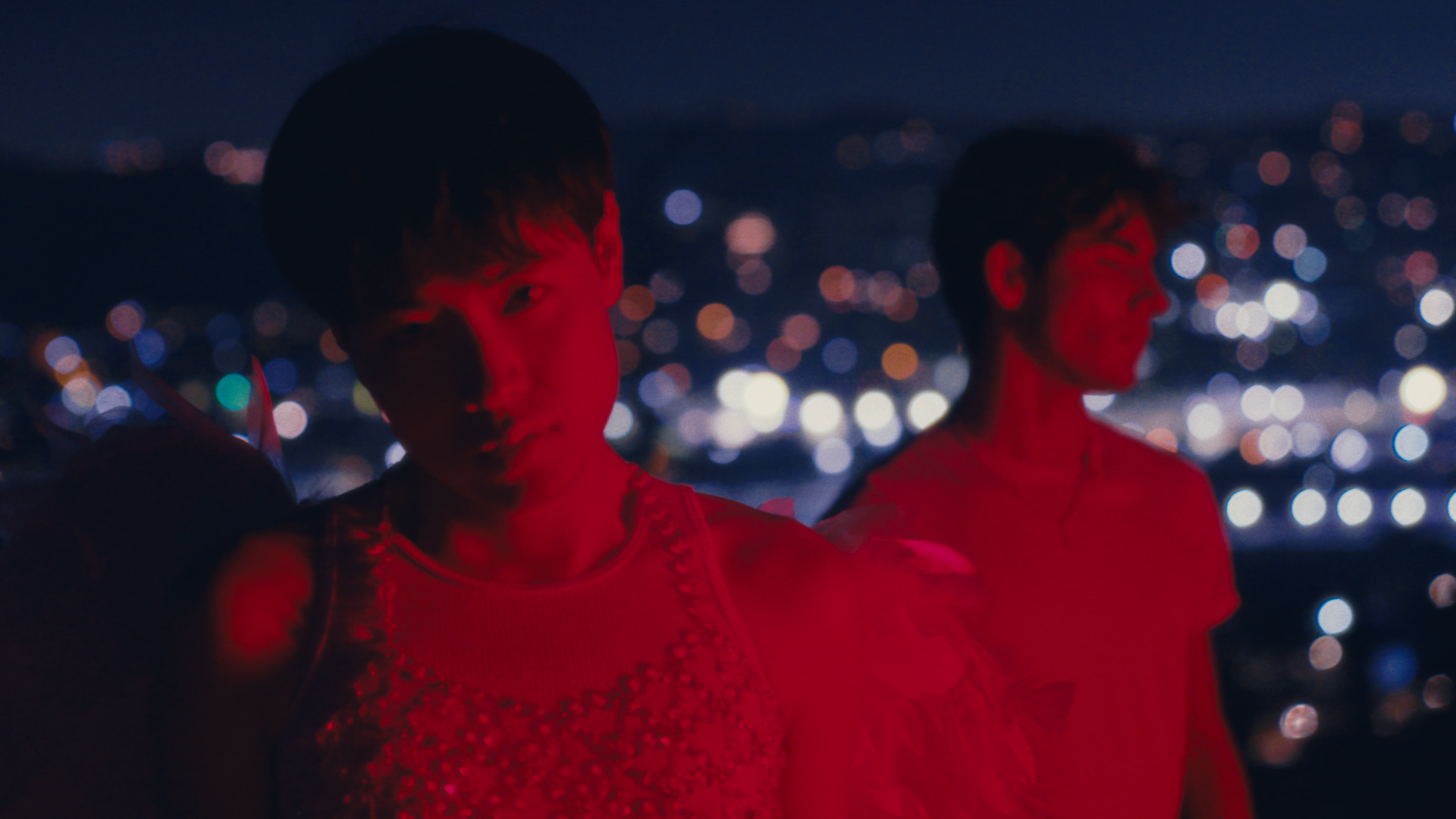 Two women stand against a cityscape at night, illuminated by bokeh lights. One woman faces forward with a serious expression, bathed in red light. The other woman is on the right, smiling and facing sideways.