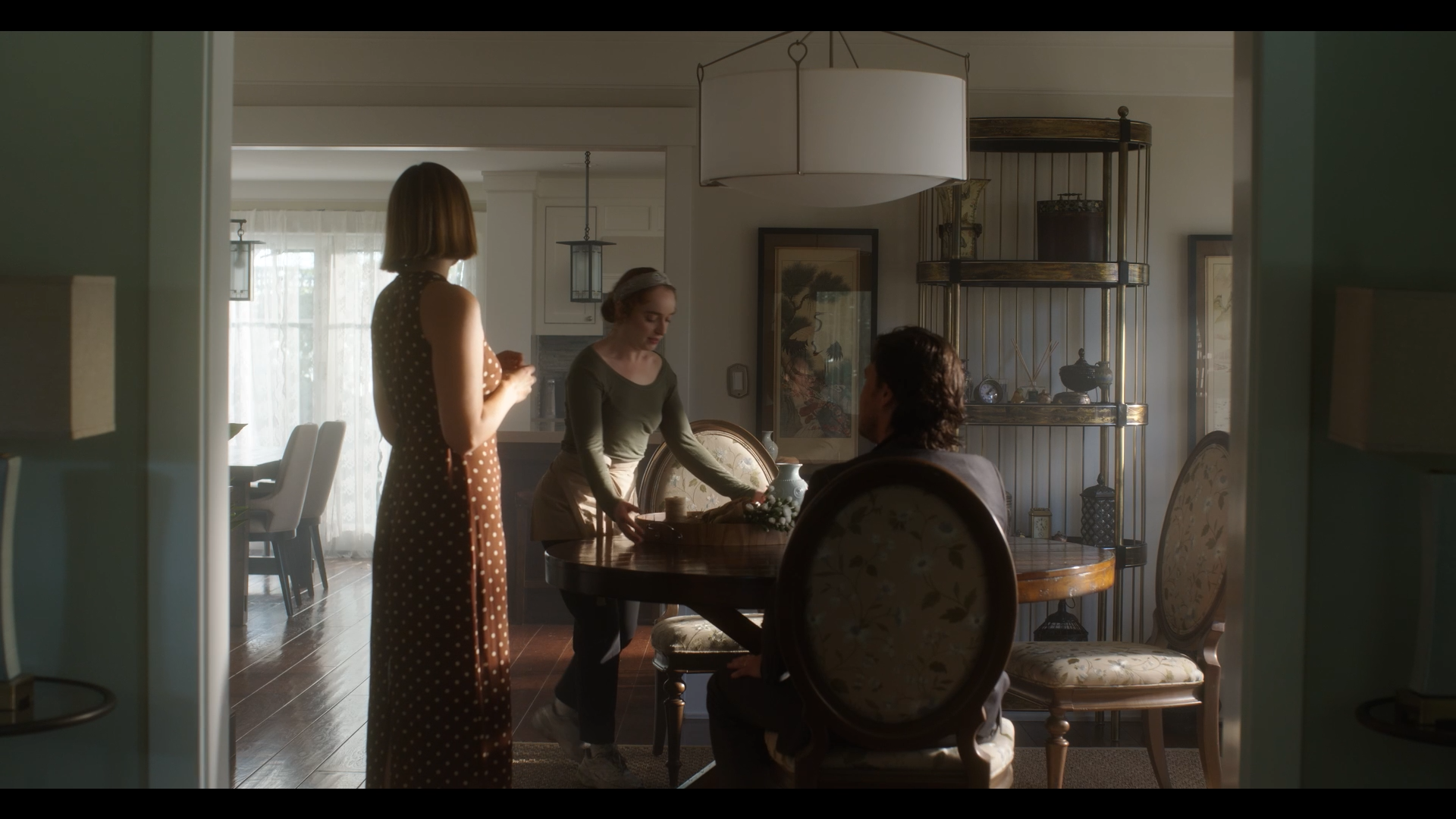 Three women are gathered in a dining area, one seated and two standing, with a table, chairs, and a shelf in the background.