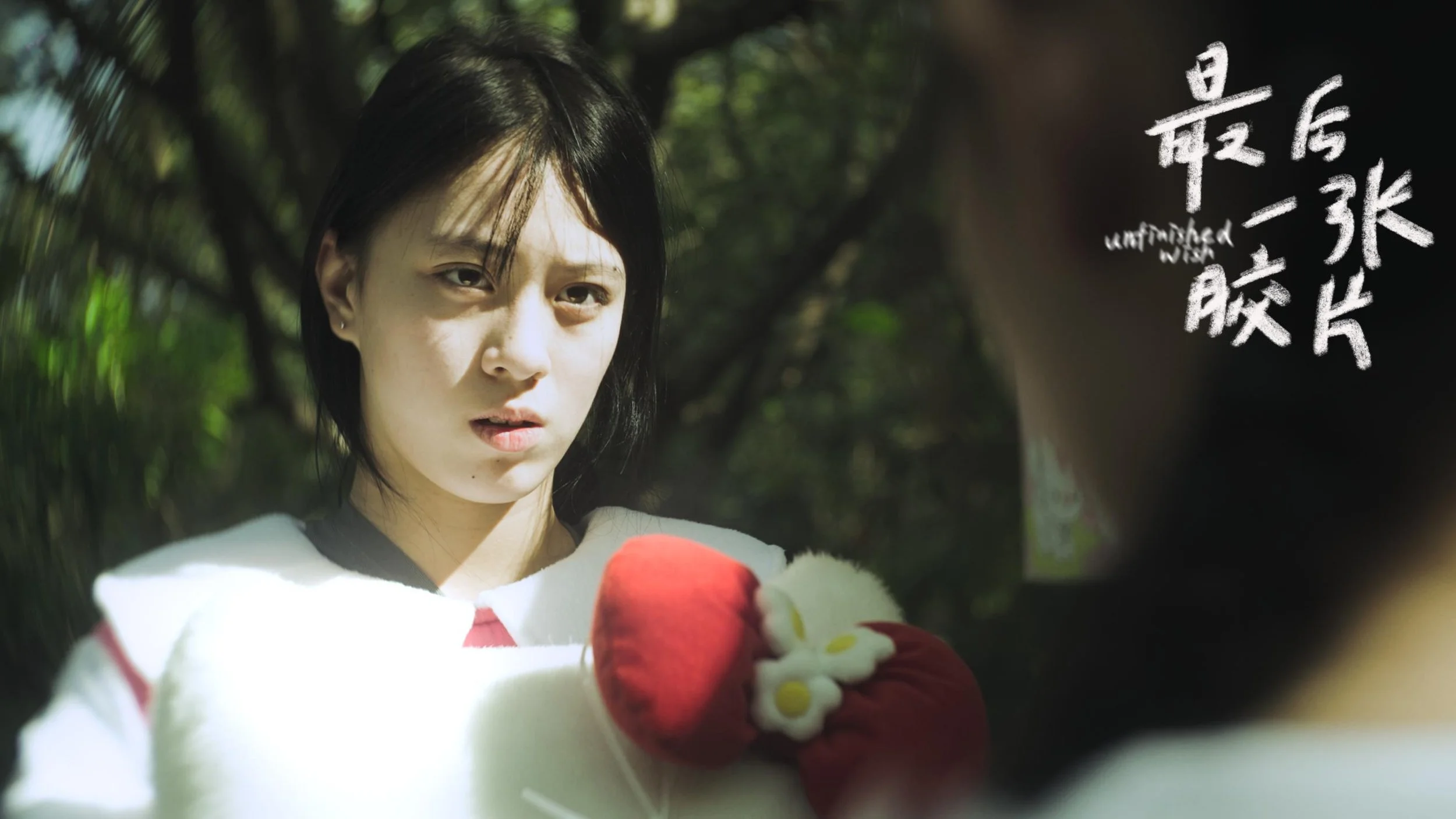 Young woman with short black hair and light skin looking thoughtfully at a plush toy held by someone outside the frame, outdoors with blurred green foliage in background, and white handwritten-style text on the right.