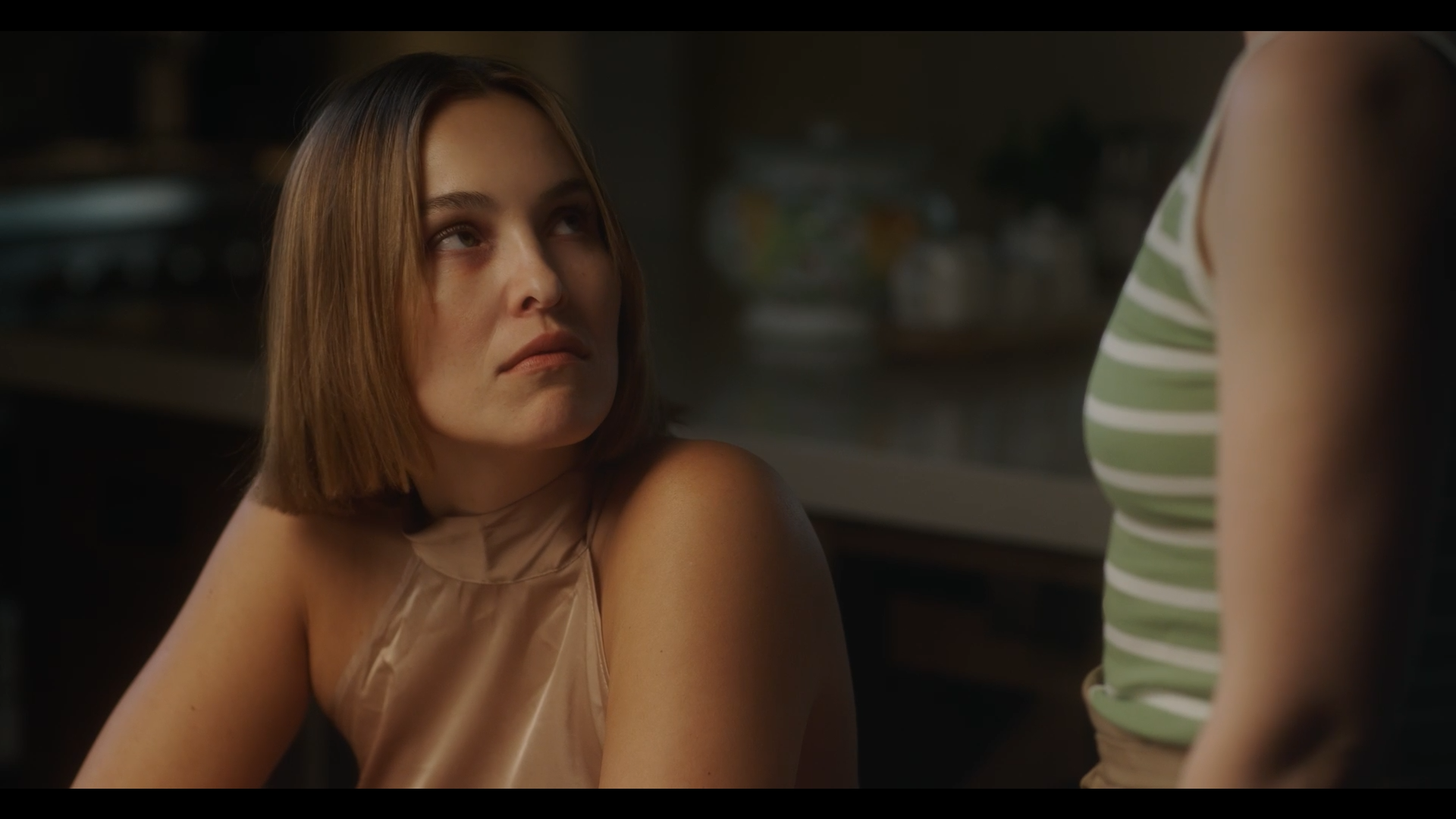 A woman with shoulder-length brown hair looking up at another person whose torso is partially visible, wearing a green and white striped sleeveless top. They are in a dimly lit indoor setting.