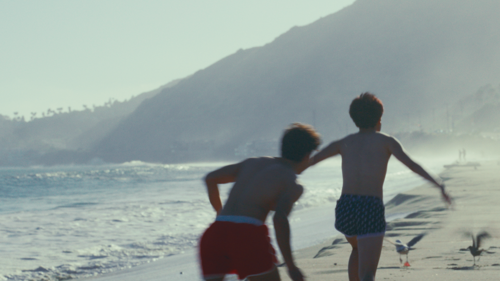Two boys running on the beach near the ocean with hills in the background, in a sunny outdoor setting.