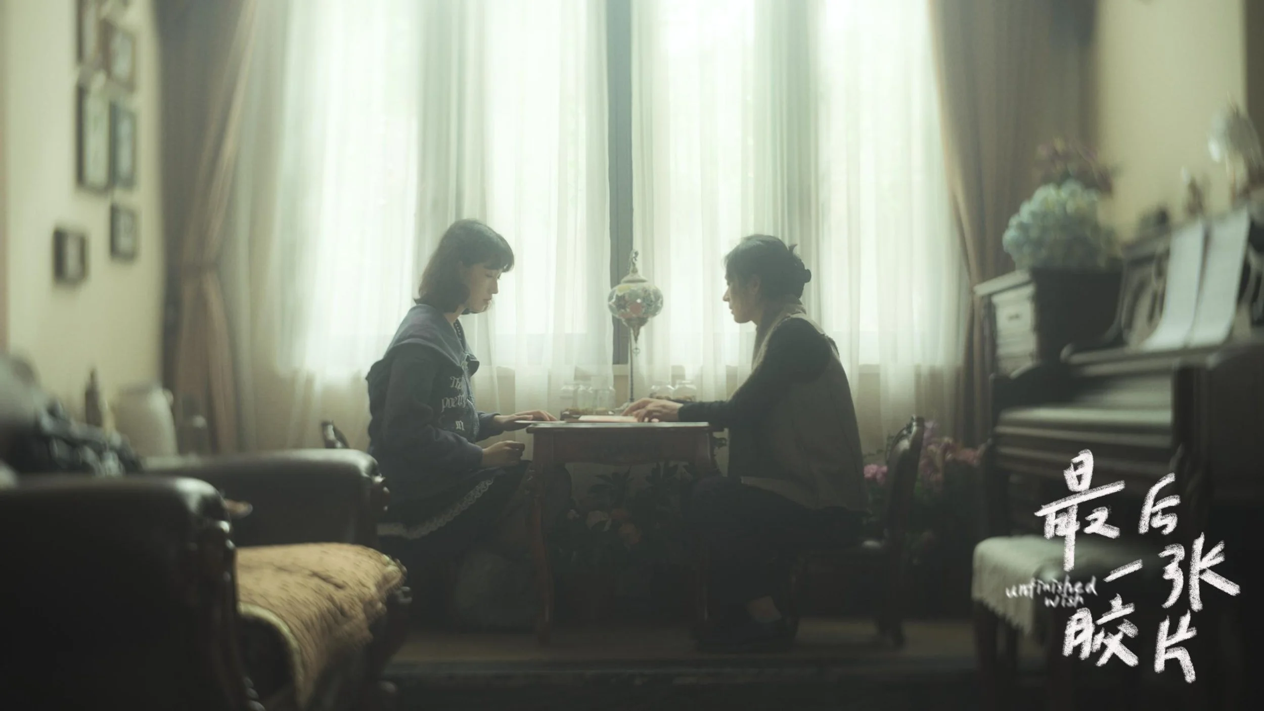 Two women sit across from each other at a small table in a well-lit room with large windows and white curtains, engaging in conversation.