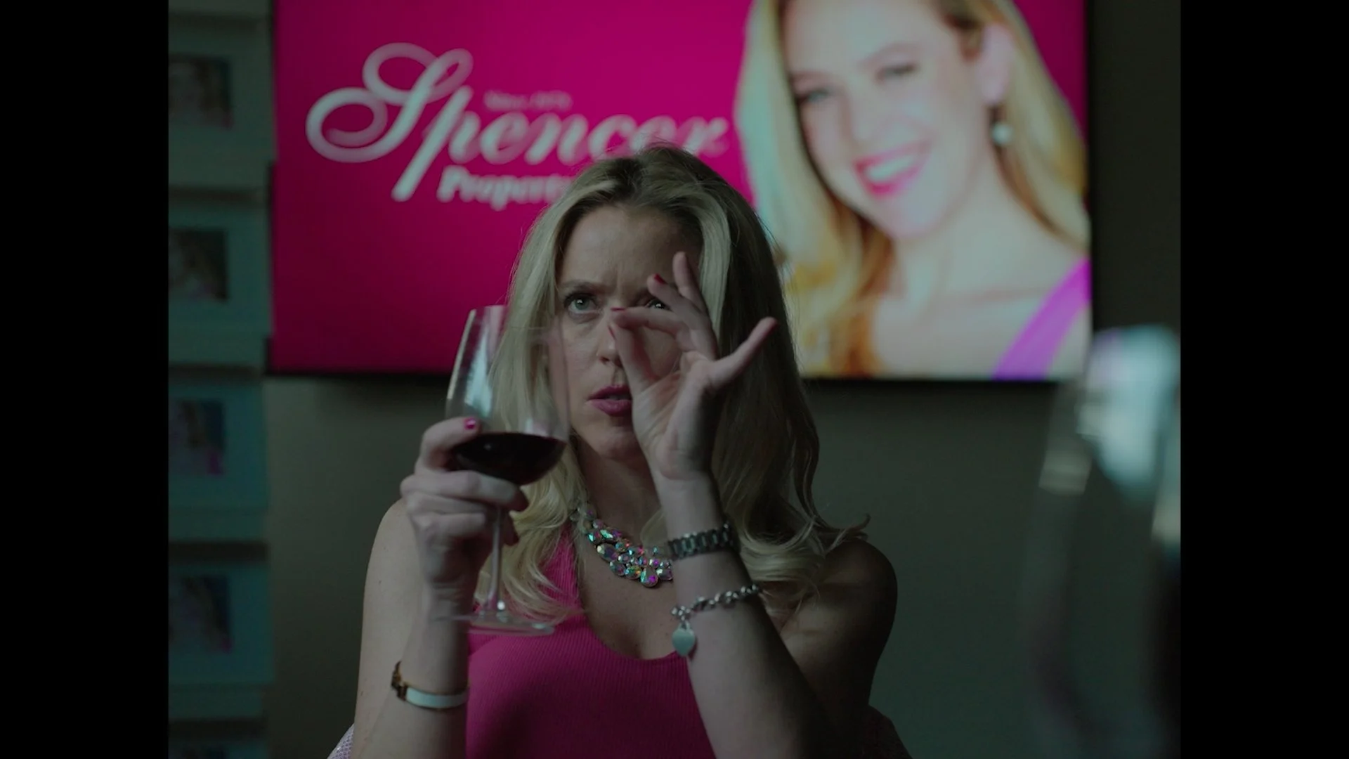 A woman with blonde hair in a pink top is holding a glass of red wine, looking through her fingers with a playful expression. Behind her, there is a bright pink advertisement poster featuring a woman with red hair.