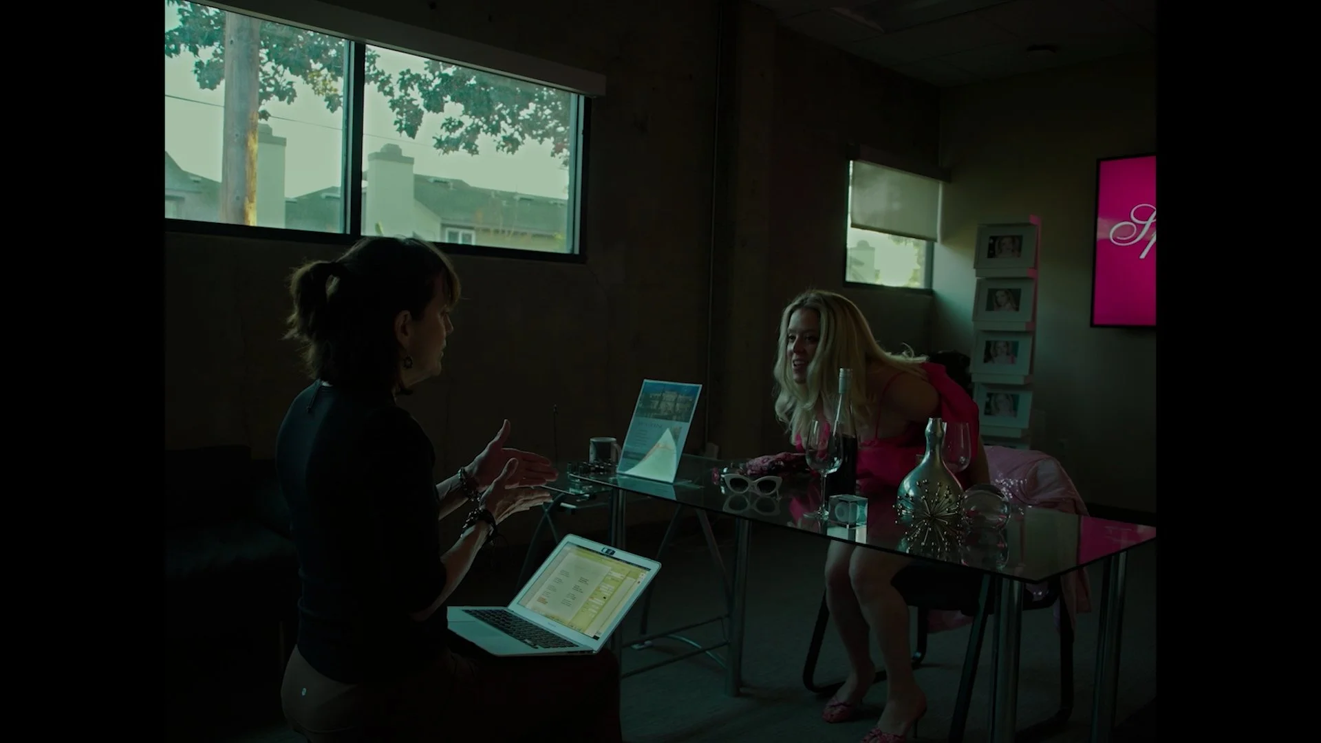 Two women having a conversation at a table inside a room with large windows, a computer, some glasses, and decorative items. One woman with dark hair is talking, while the other with blonde hair is leaning on the table. The room has natural light com