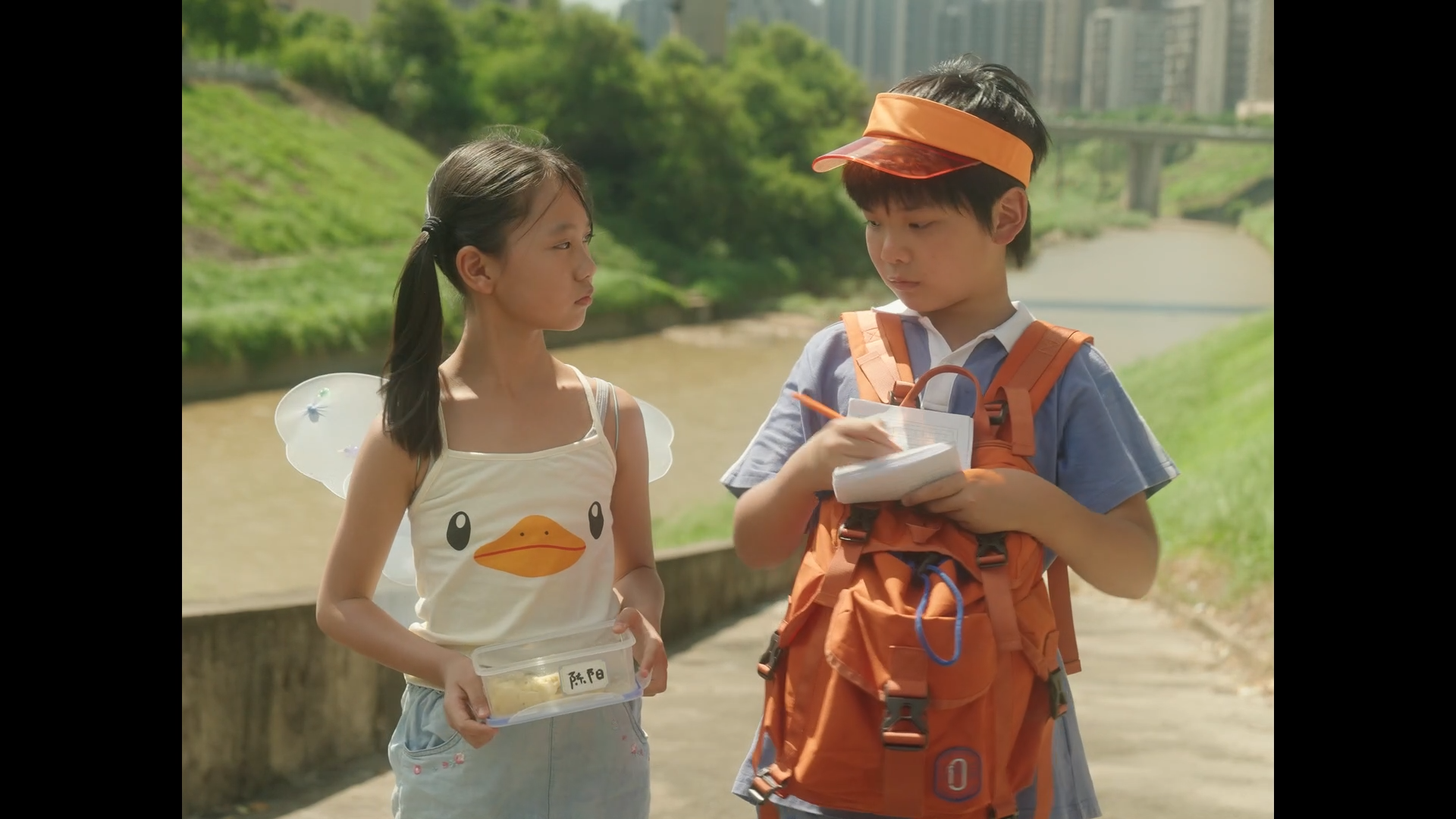 A young girl with fairy wings and a duck design on her tank top is holding a container of food, standing outdoors by a river. A boy with a backpack and visor is writing in a notebook and talking to her. Green trees, a river, and city buildings are vi