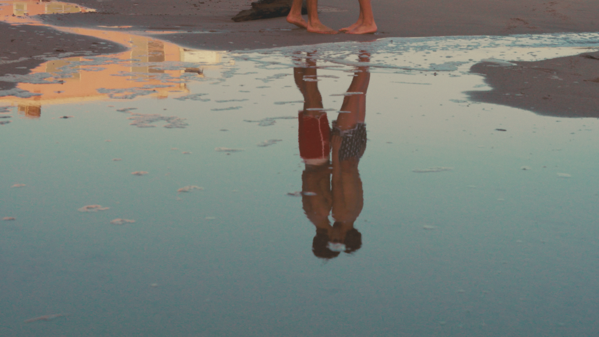 Two children standing on wet sand at the beach, reflected in a large puddle, holding hands, with the reflection showing a clear view of their legs and crests.