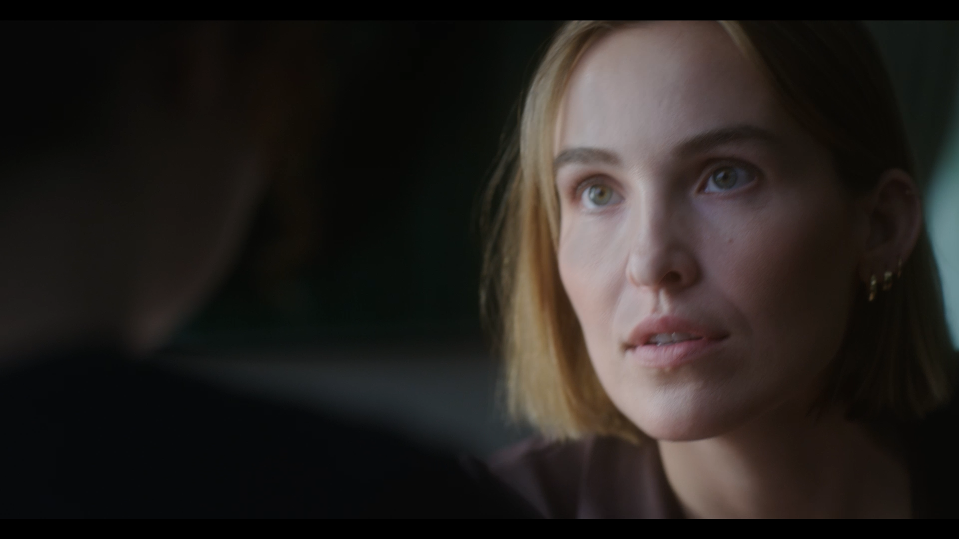 Close-up of a woman with blonde hair and piercing eyes, looking intently at someone or something off-camera.
