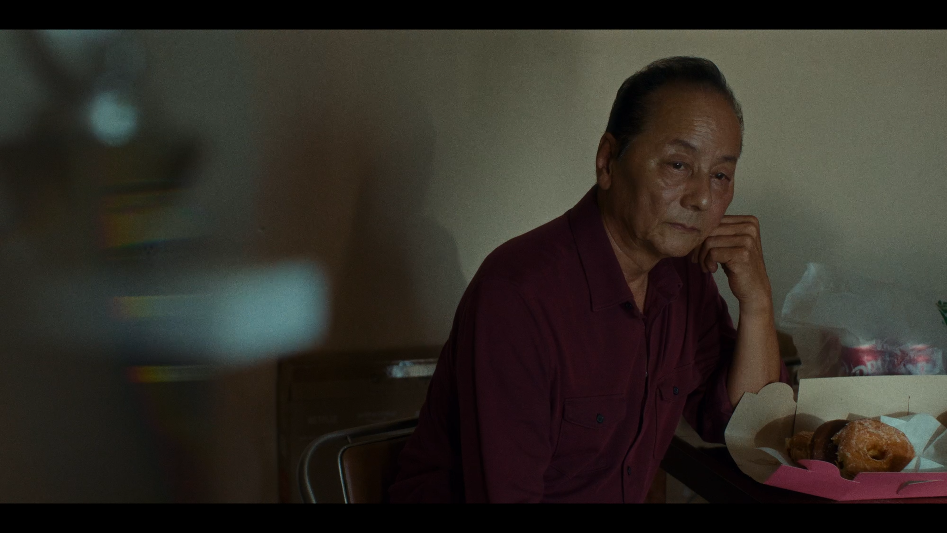 An older man with dark hair and a contemplative expression, sitting at a table with a box of donuts and a pink napkin, in a dimly lit room.