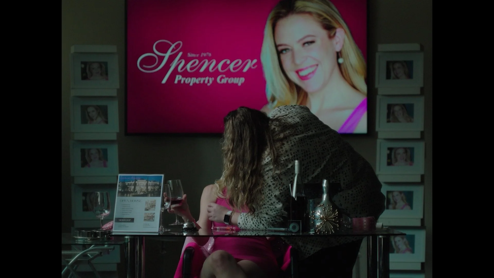 A woman with long dark hair leaning over a young girl with blonde hair, seated at a table with wine glasses and a menu. Behind them, a large pink sign reads 'Since 1978 Spencer Property Group' with a portrait of a woman.