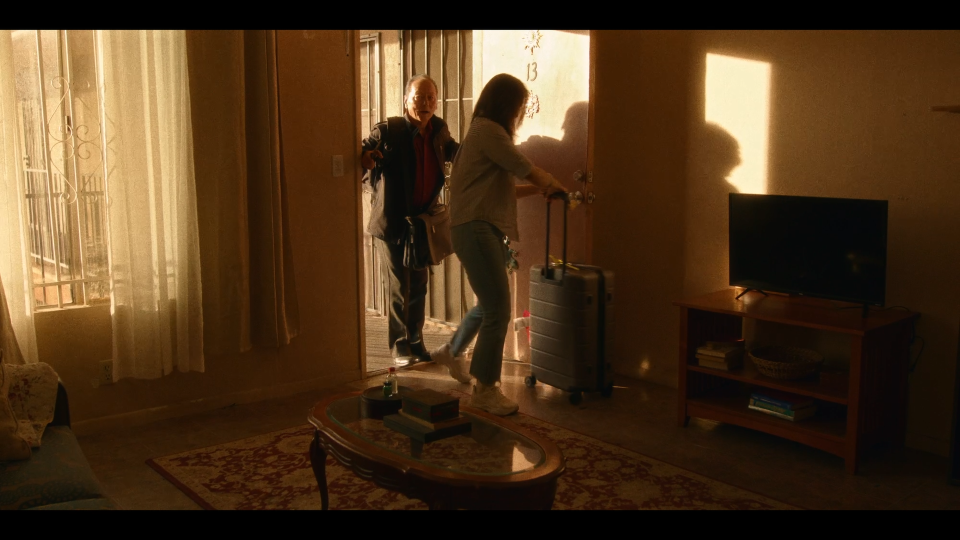A woman with a rolling suitcase entering a home with an older man welcoming her at the door. The interior is dimly lit with sunlight streaming in, revealing a television and a bookshelf.