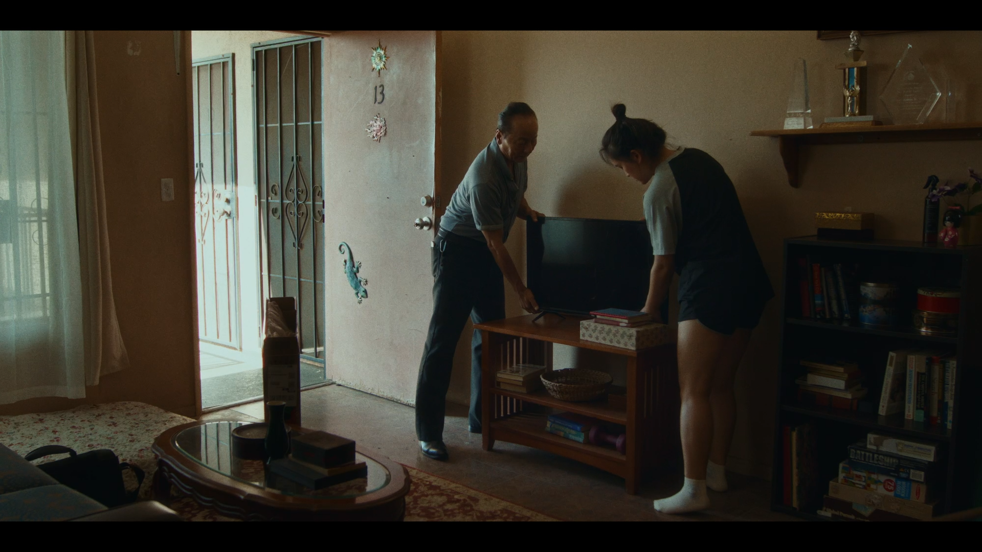 Two people, an older man and a young woman, are standing in a living room near the front door, looking at a small television on a wooden stand. The young woman is wearing a black and white top with shorts and white socks, while the older man is dress