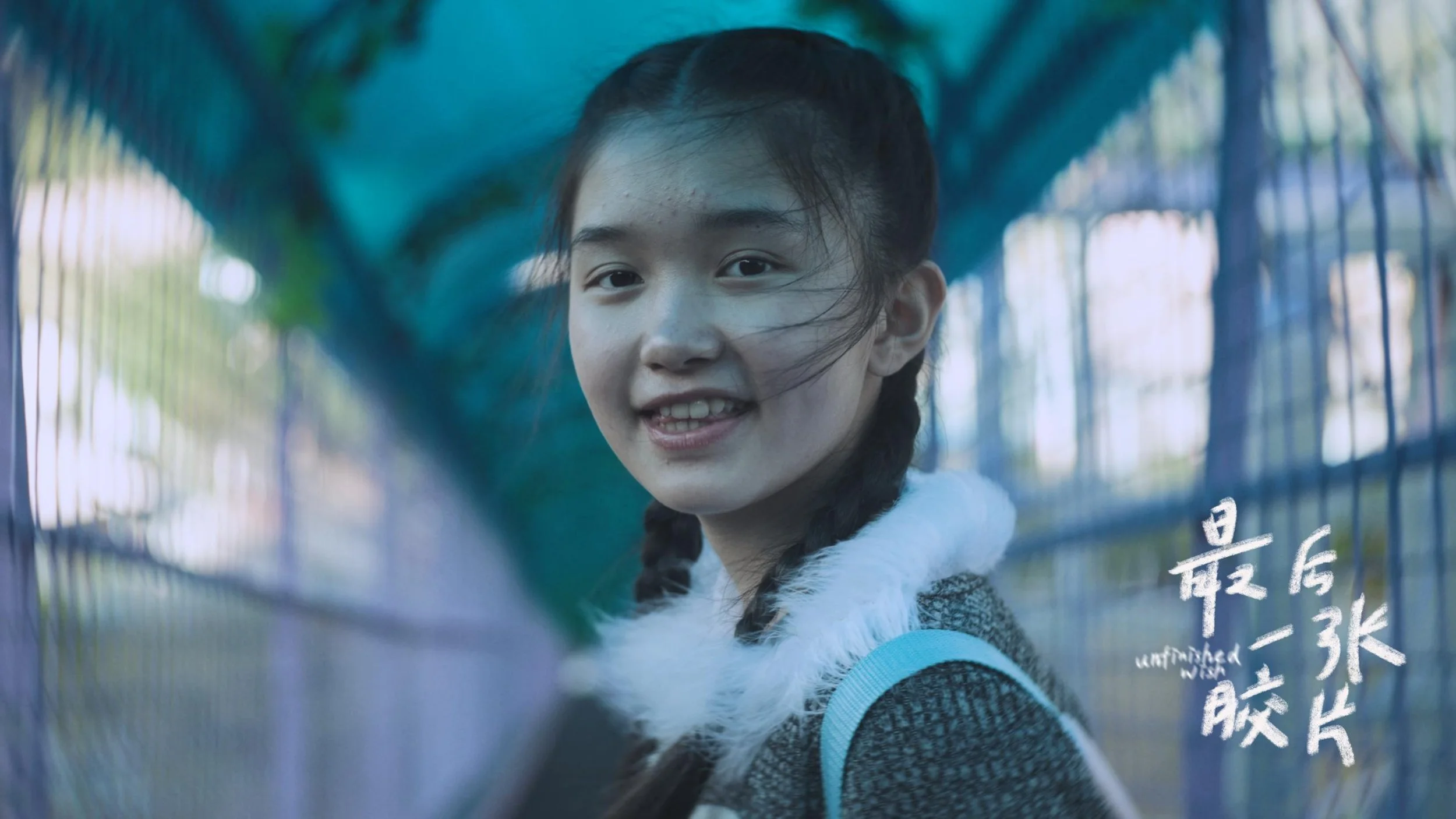 A young girl with braided hair smiling behind a glass surface with reflective writing in a foreign language.