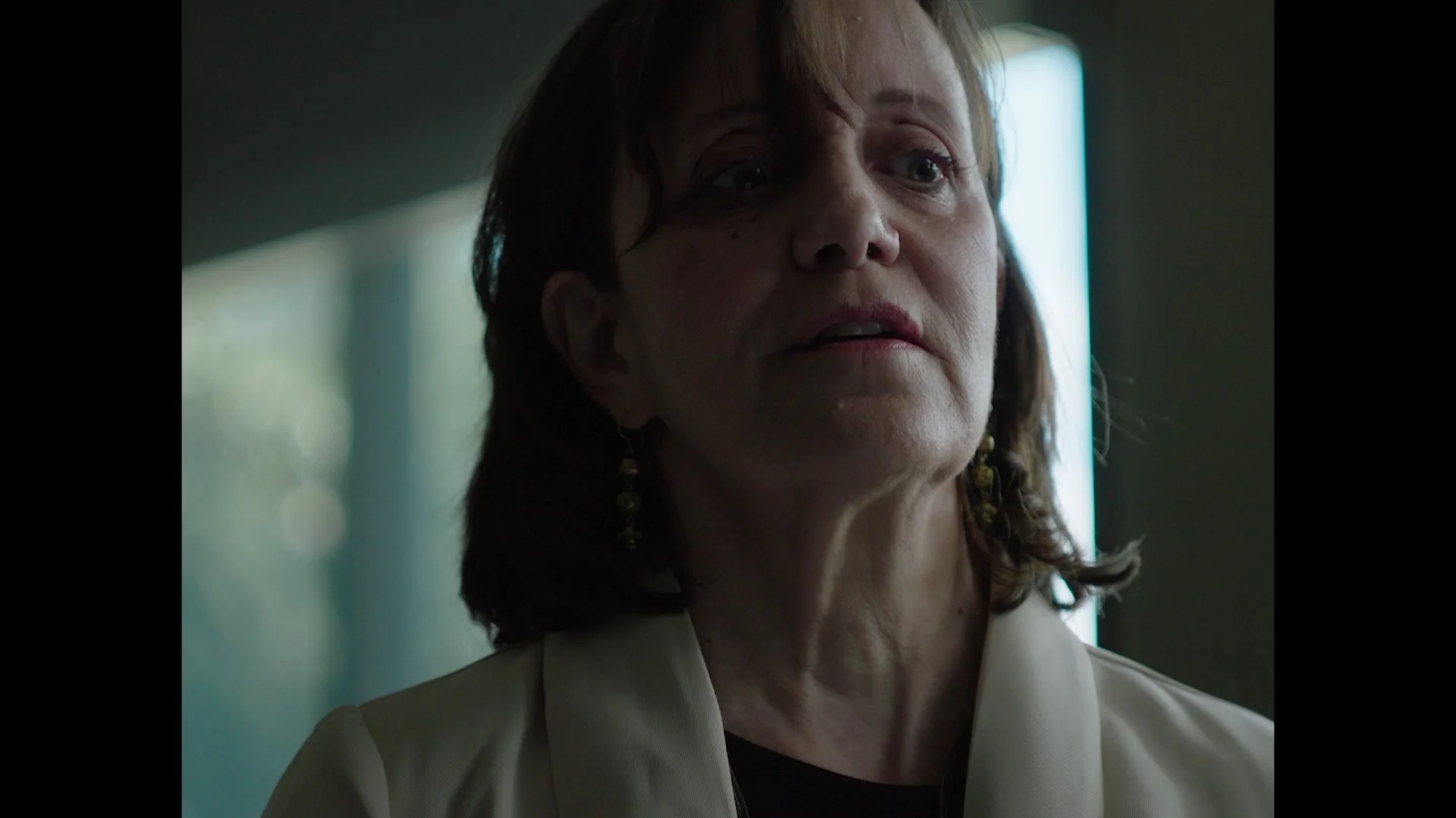 Close-up of a middle-aged woman with reddish-brown hair, wearing a beige blazer and earrings, looking slightly to the side in a dimly lit room with a bright vertical light in the background.