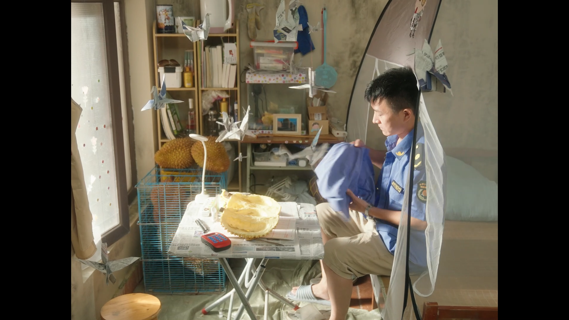 A young man in uniform is sitting at a table in a room, folding a cloth. The table has a cake, newspaper, and a calculator on it. There are origami paper cranes hanging in front of him, a cage with chickens, and bookshelves in the background. Sunligh