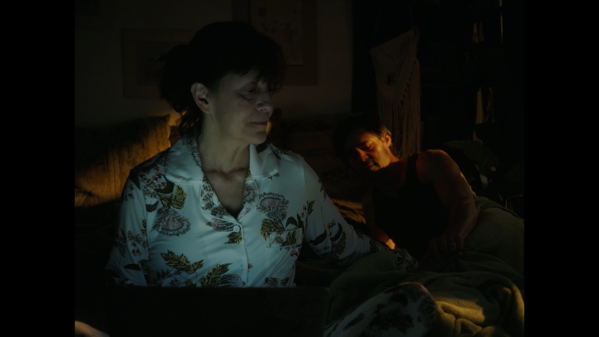 A woman with short dark hair wearing a floral button-up shirt sitting on a couch in a dimly lit room, with another woman with short hair and sleeveless top resting behind her.