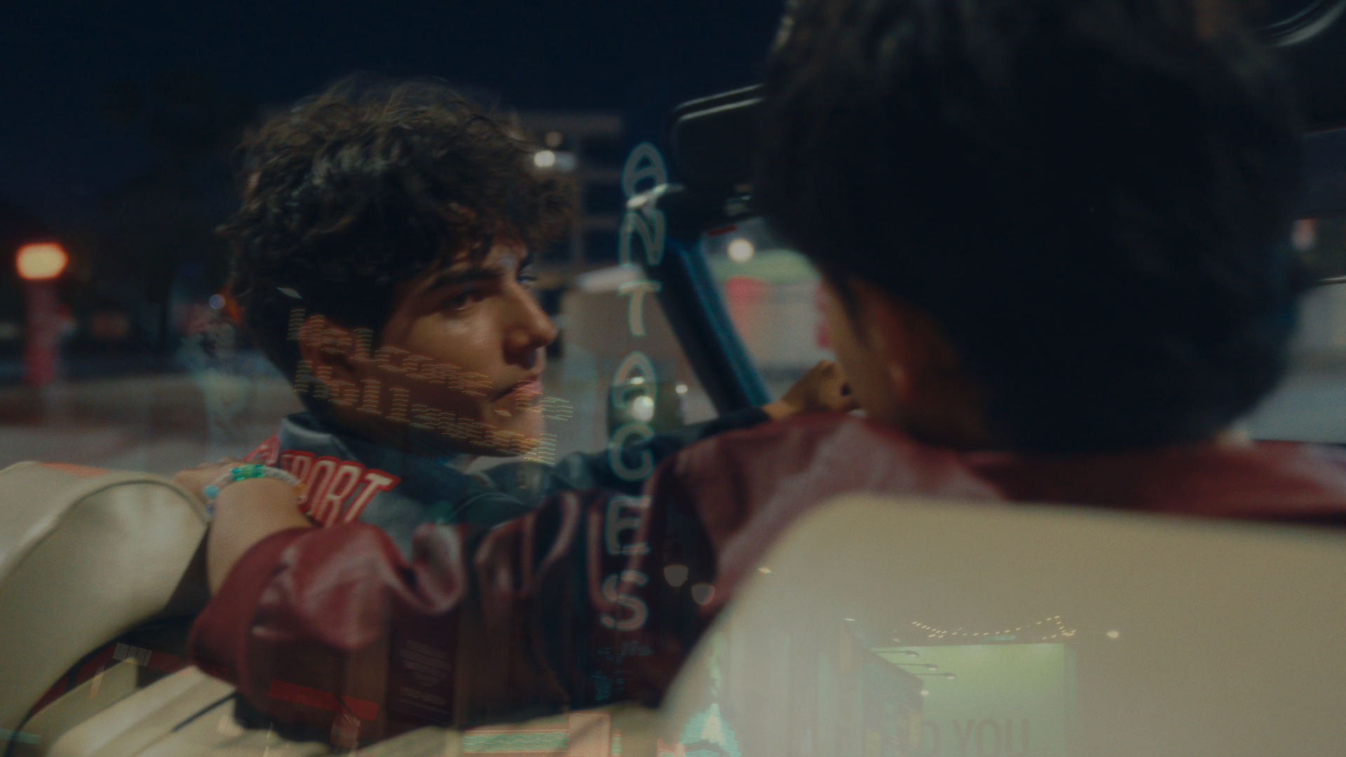 Two young men sitting inside a car, viewed through the window at night, engaged in a conversation with city lights reflected on the glass.