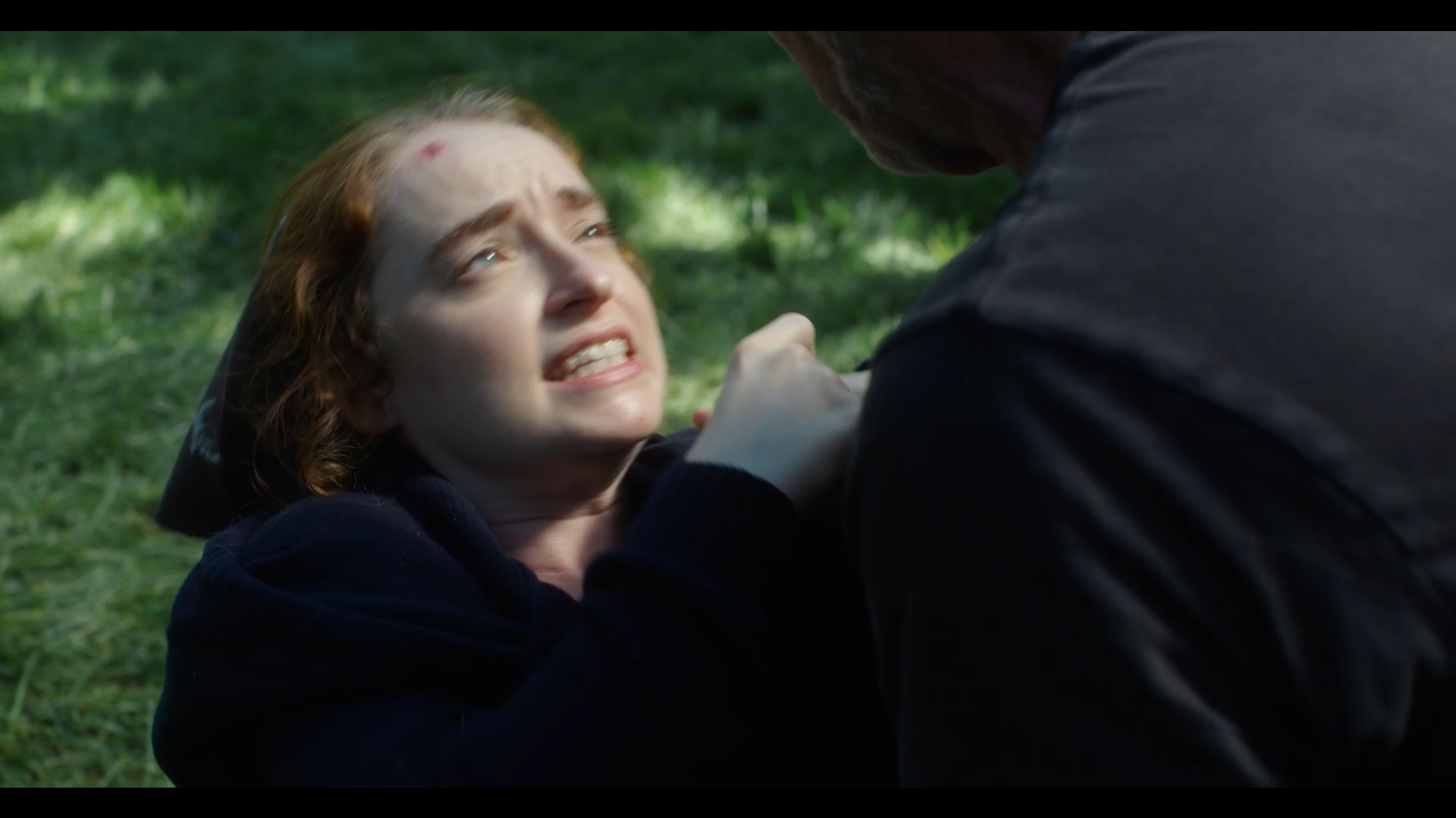 A woman with red hair and a bruise on her forehead is on the ground, looking distressed and crying. A man, whose face is not visible, is leaning over her, grasping her arm in a tense manner.