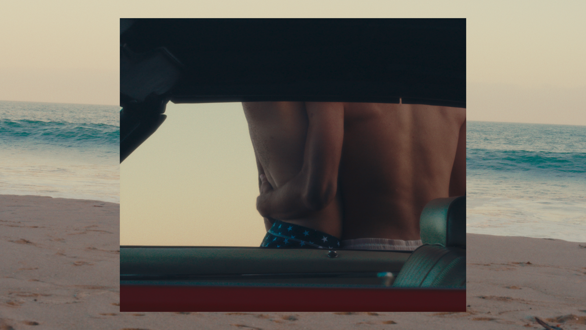 People at the beach viewed through a car window, showing a person holding a surfboard and another person standing nearby with the ocean in the background.