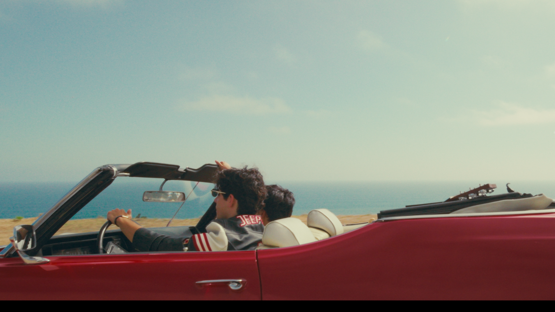A red convertible car driving along a coastal road with two young men inside, one wearing sunglasses and a jacket with 'JEEP' on the sleeve, with the ocean and sky in the background.