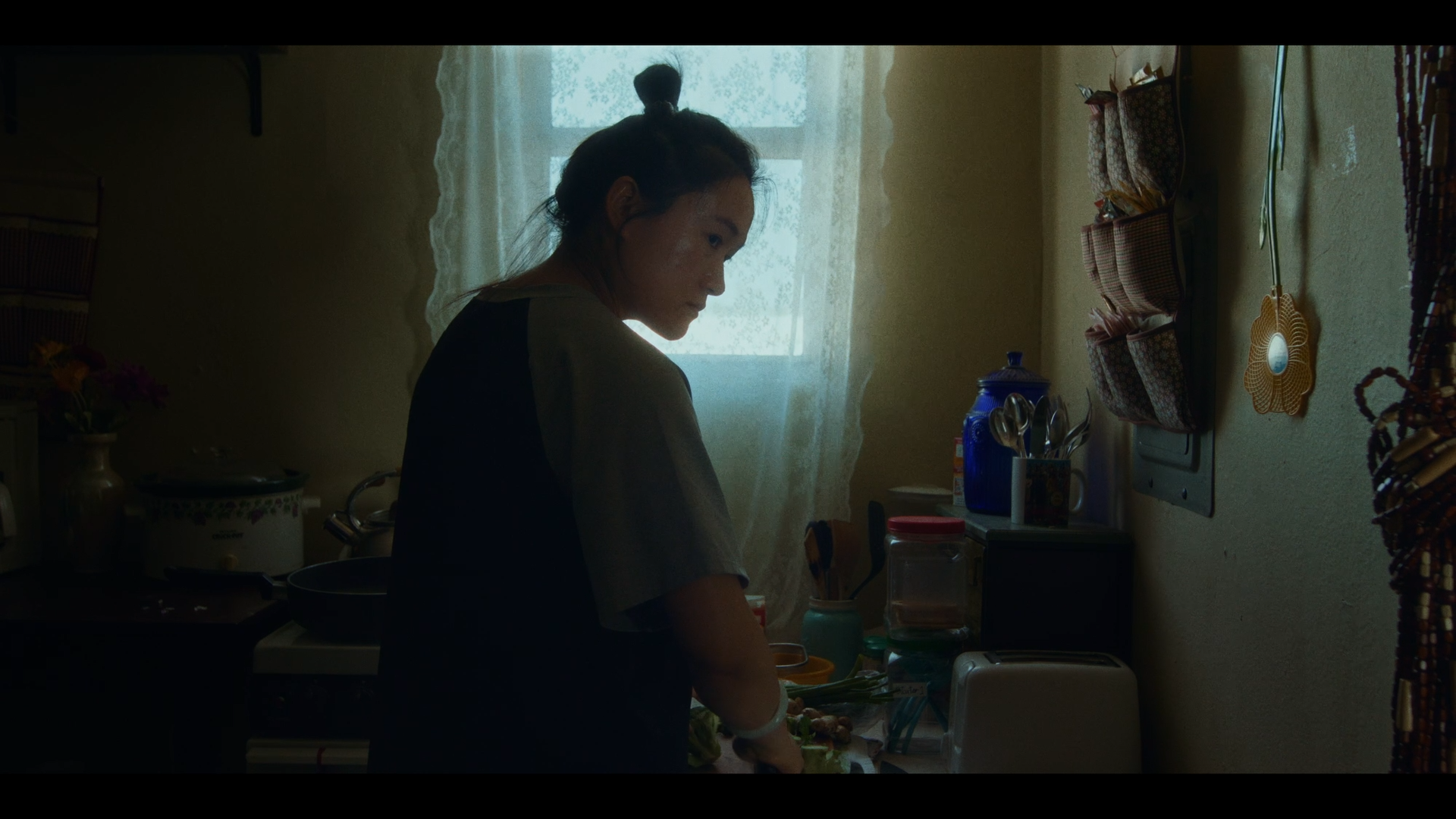 A woman with her hair in a top knot preparing food on a kitchen counter in a dimly lit kitchen with a window and hanging storage on the wall.