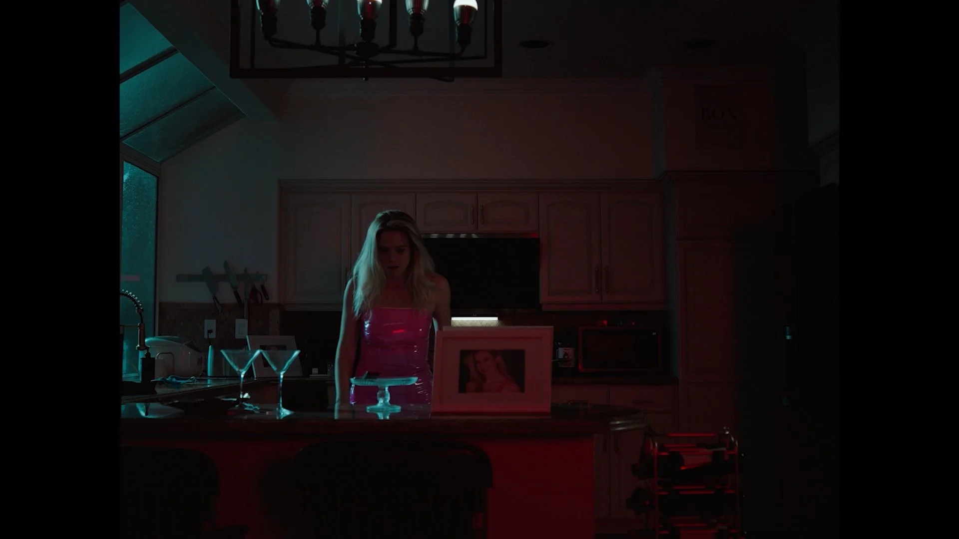 A woman with blonde hair in a pink dress standing behind a kitchen counter at night, with two martini glasses and a framed photo on the counter, via dim lighting.