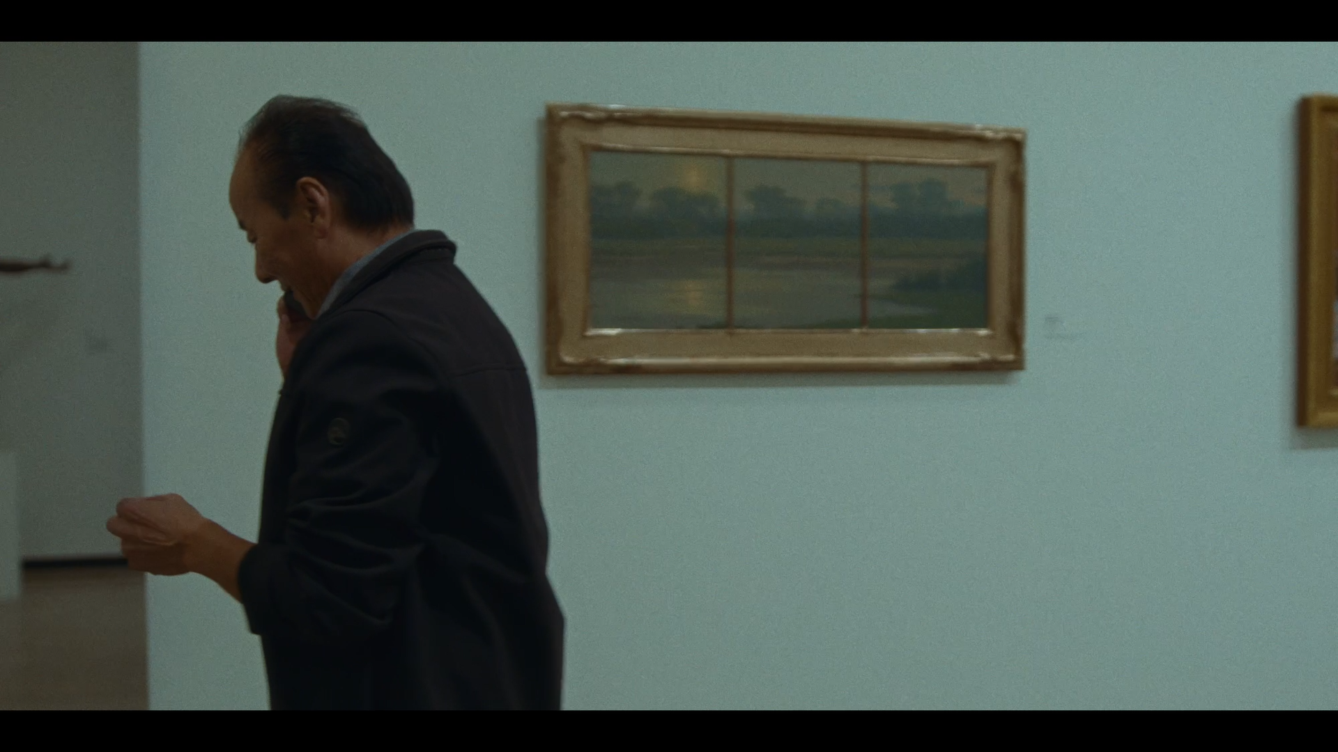 A man standing and talking on a cellphone in a room with light green walls, with three framed landscape paintings on the wall.