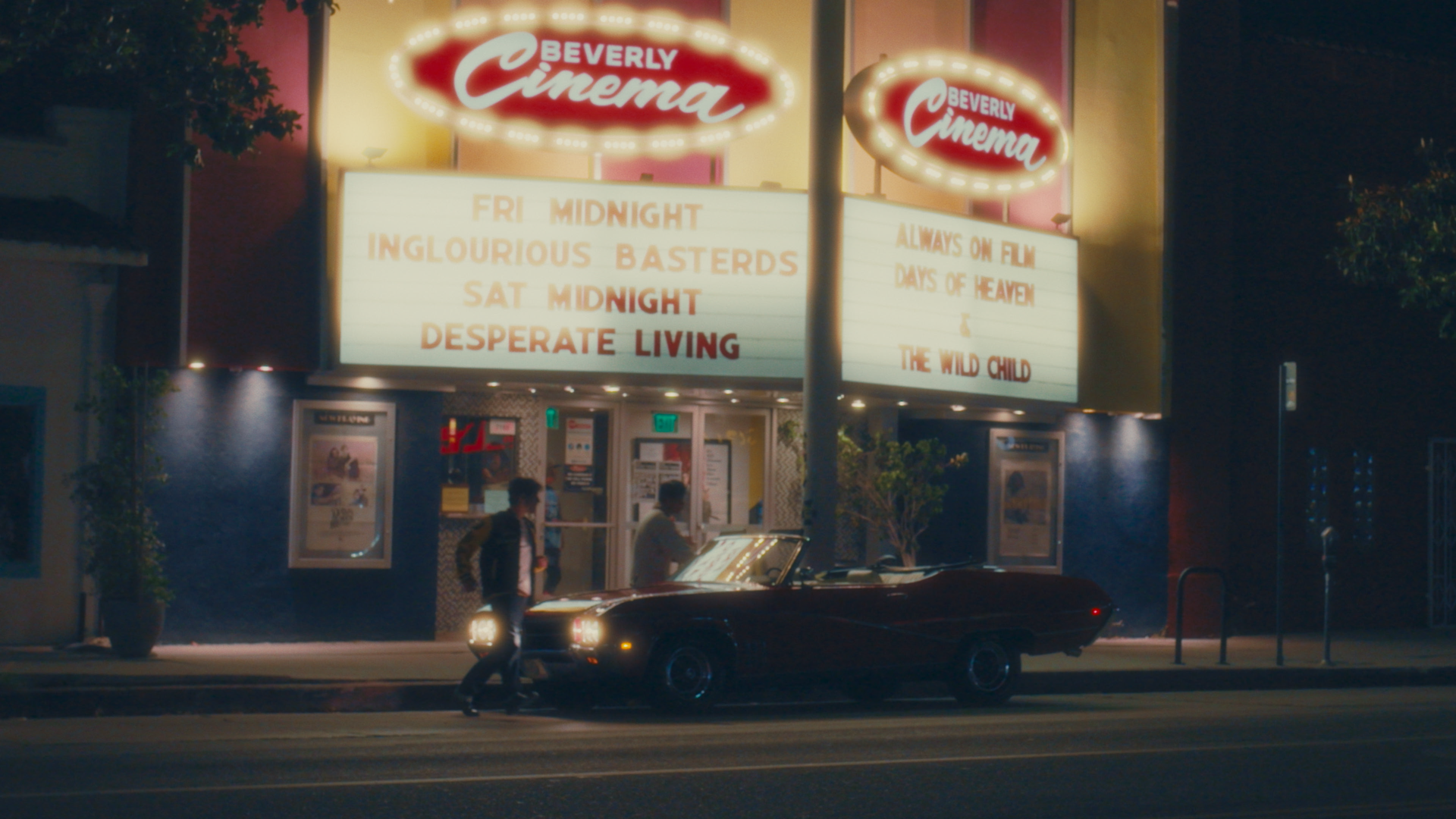 Nighttime scene outside Beverly Cinema with illuminated marquee listing horror movies and showtimes, a vintage black convertible car parked near the entrance, and two men walking past.
