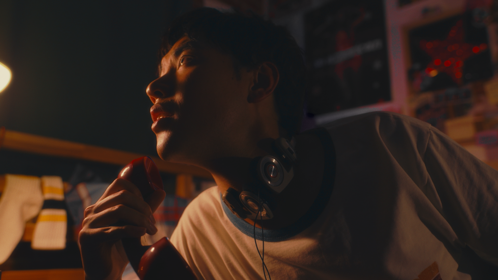 A young man with headphones around his neck holding a red object in a dimly lit room with colorful shelves and posters in the background.