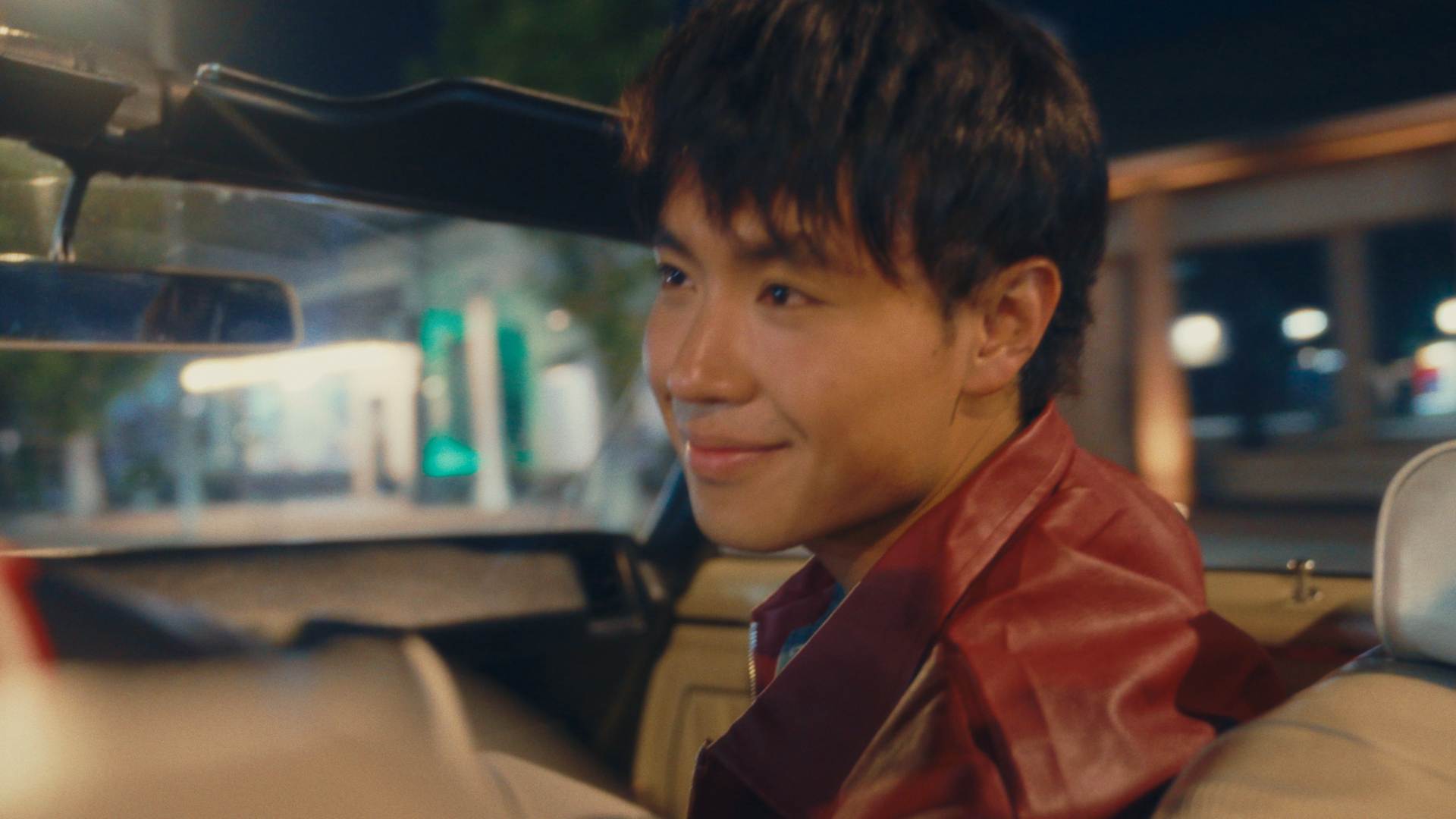 A young man with dark hair and a slight smile, sitting in the backseat of a car during nighttime or evening, wearing a red leather jacket.