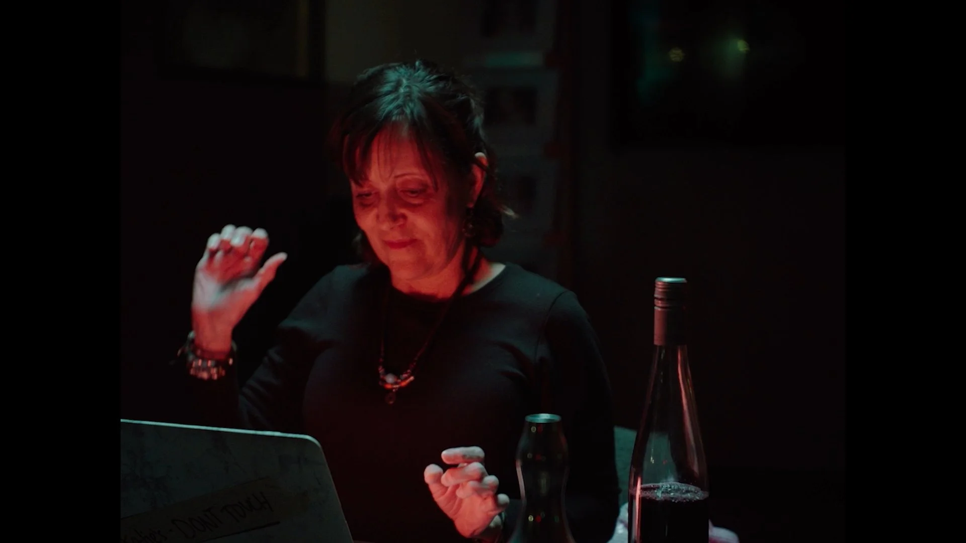 A woman with short dark hair, wearing a black shirt and colorful jewelry, appears to be in a dimly lit setting, possibly a bar or restaurant, illuminated by red lighting. She is looking at her phone with a slight smile, with a bottle and glass on the