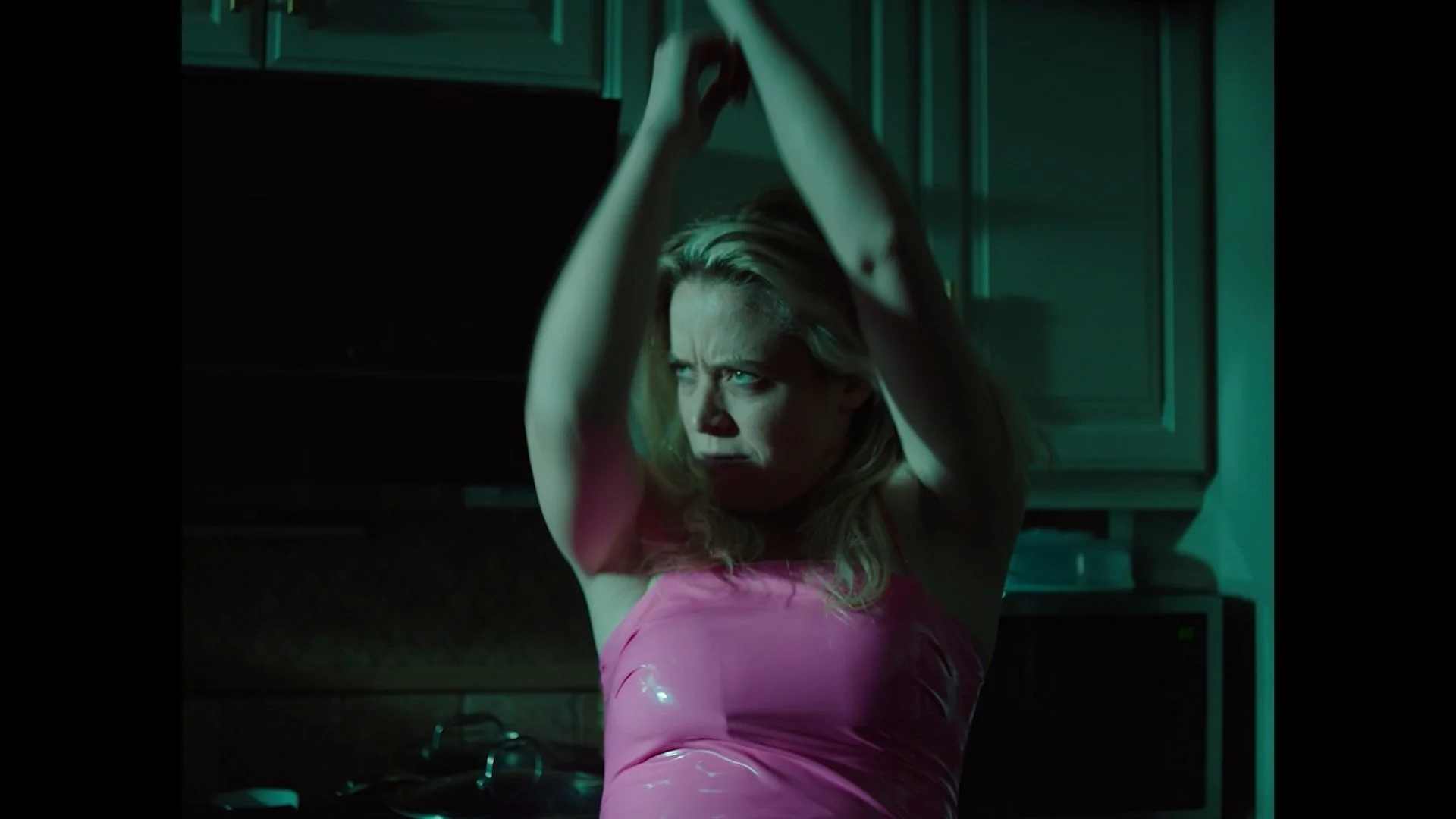 A woman with blonde hair wearing a pink top, standing with her arms raised above her head in a room with dark and greenish lighting.
