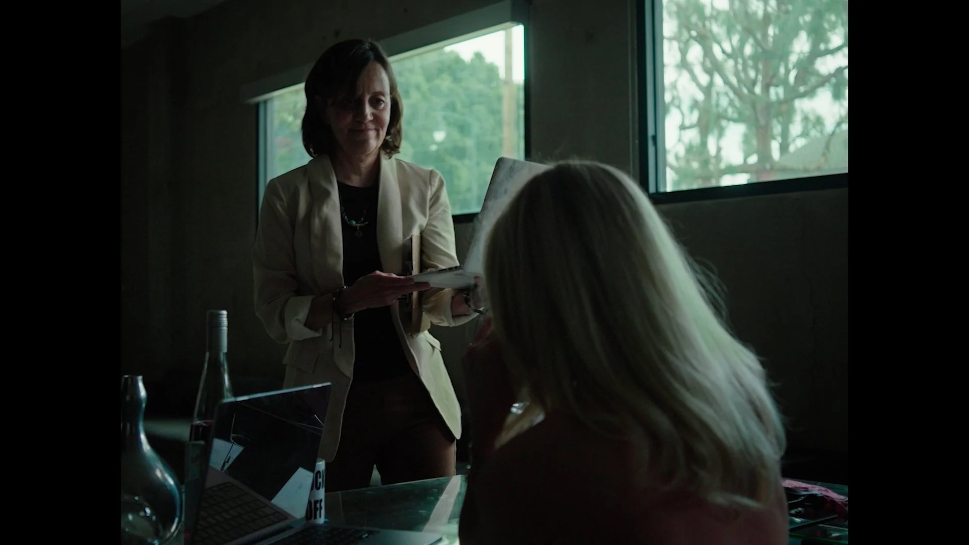 A woman with short dark hair standing and smiling while holding a tablet, facing a seated person with long blonde hair in a room with large windows showing trees outside.